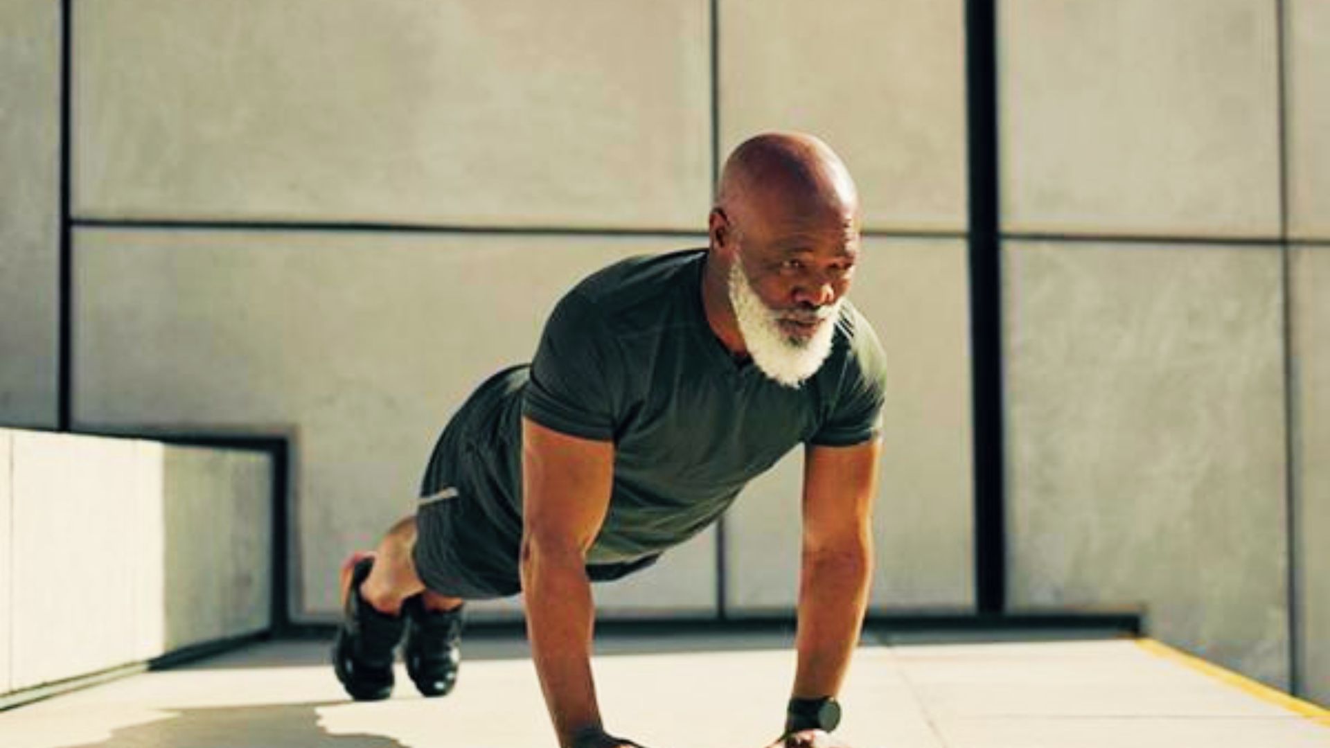 An older man with a white beard holds a strong plank position on concrete outdoors in direct sunlight.