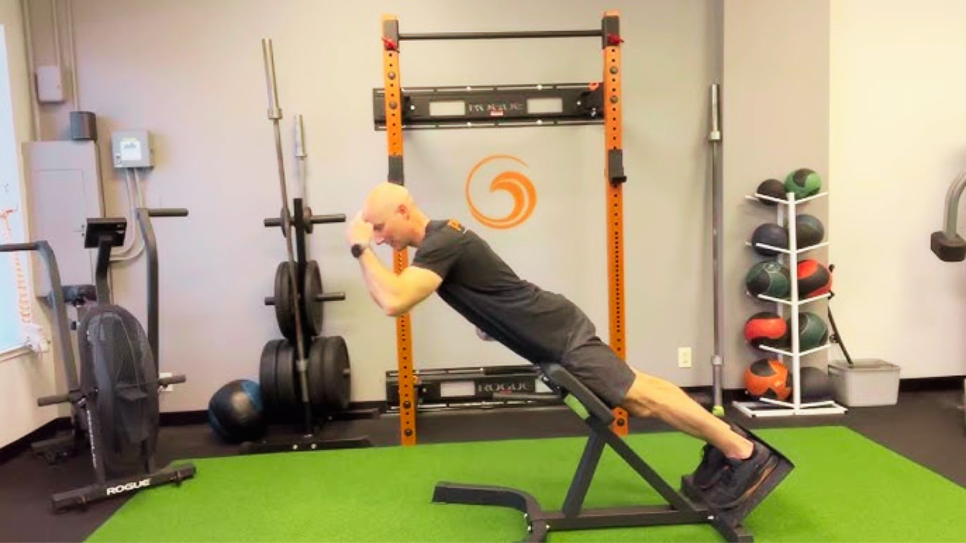 A bald man in black athletic wear performs a bodyweight back extension on a 45-degree Roman chair.