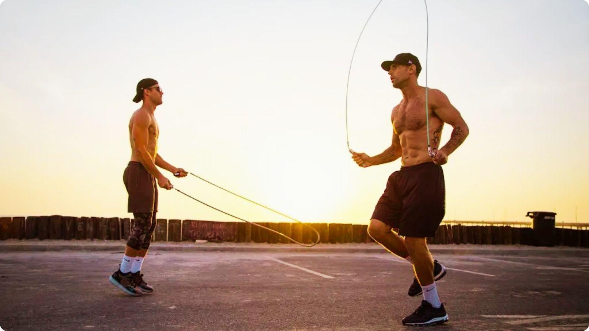 Two men jump rope shirtless on pavement during golden hour near the ocean
