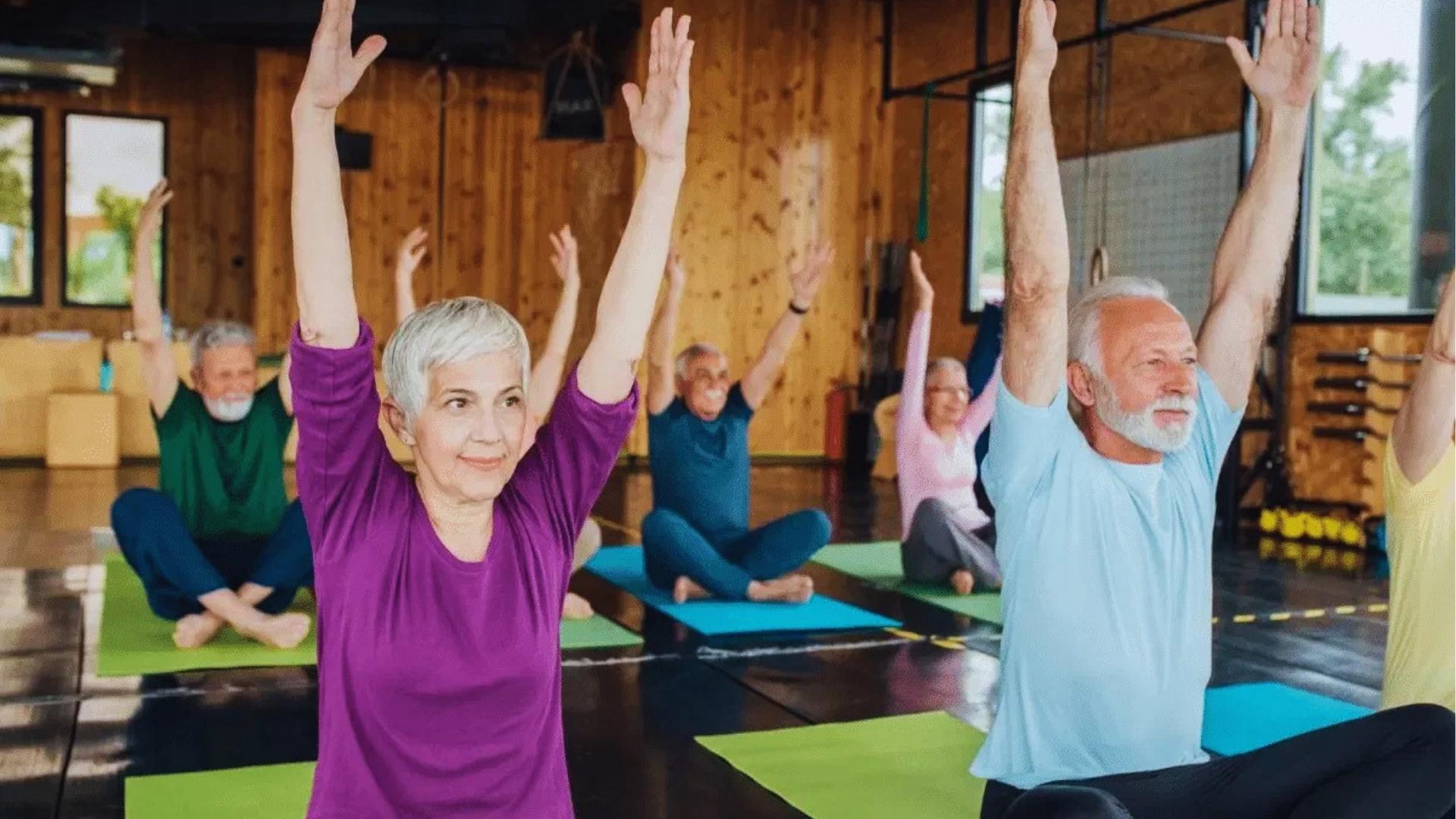 Group of seniors in colorful shirts sits on yoga mats with arms raised during gentle chair yoga class.