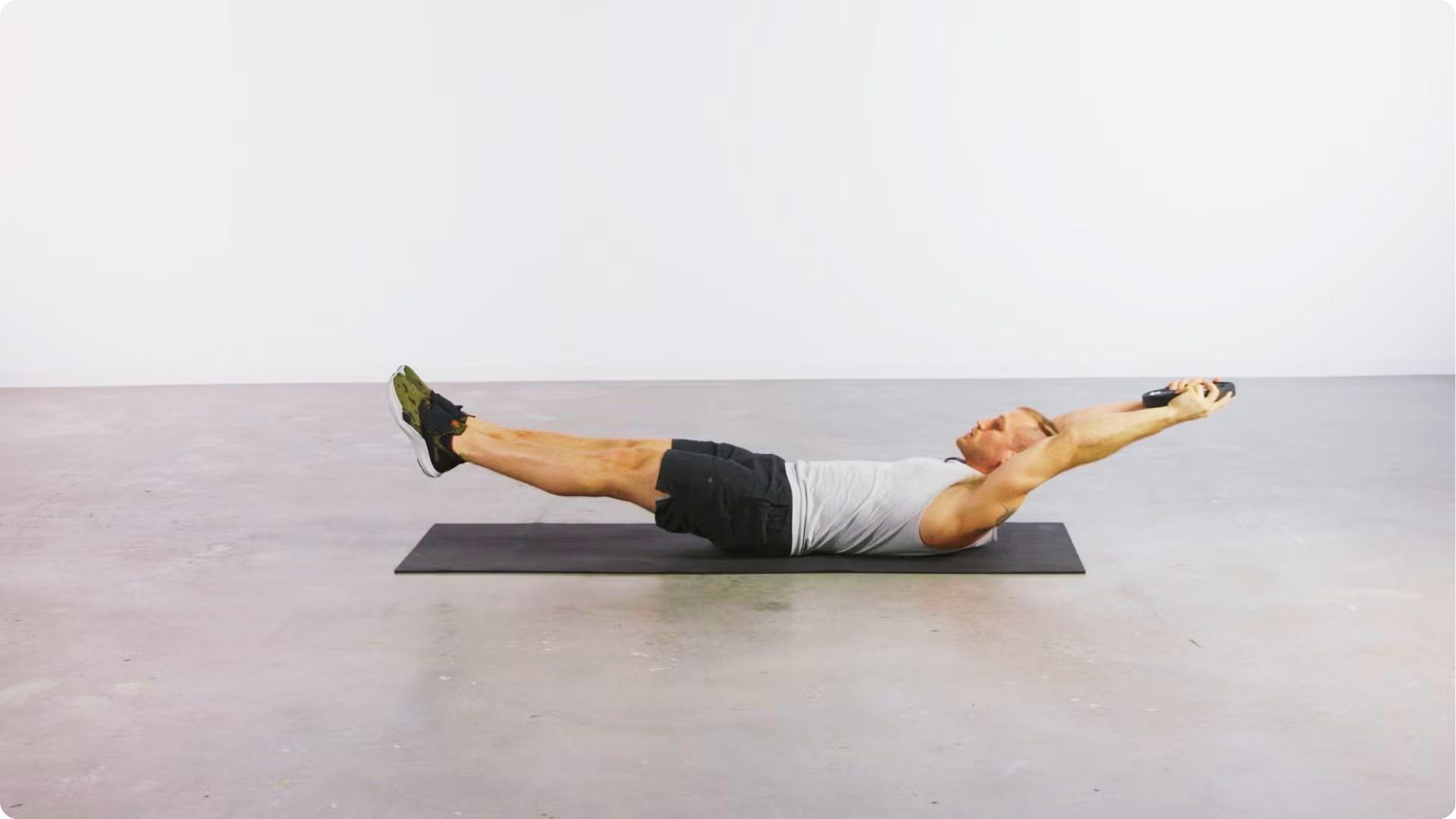 A man in a white tank top holds a weighted hollow body hold on a mat in a minimal studio.