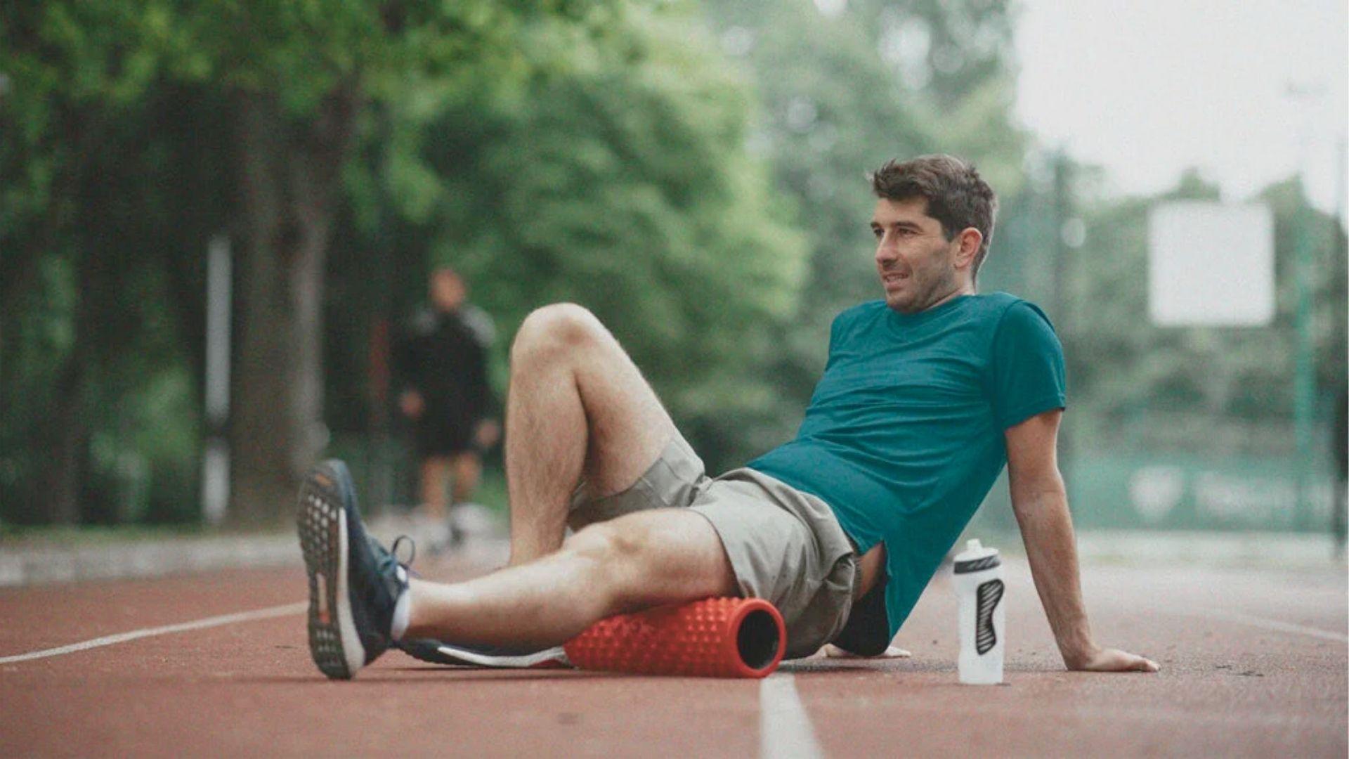Man in teal shirt sits on a track using a red foam roller on his hamstring with a water bottle nearby.