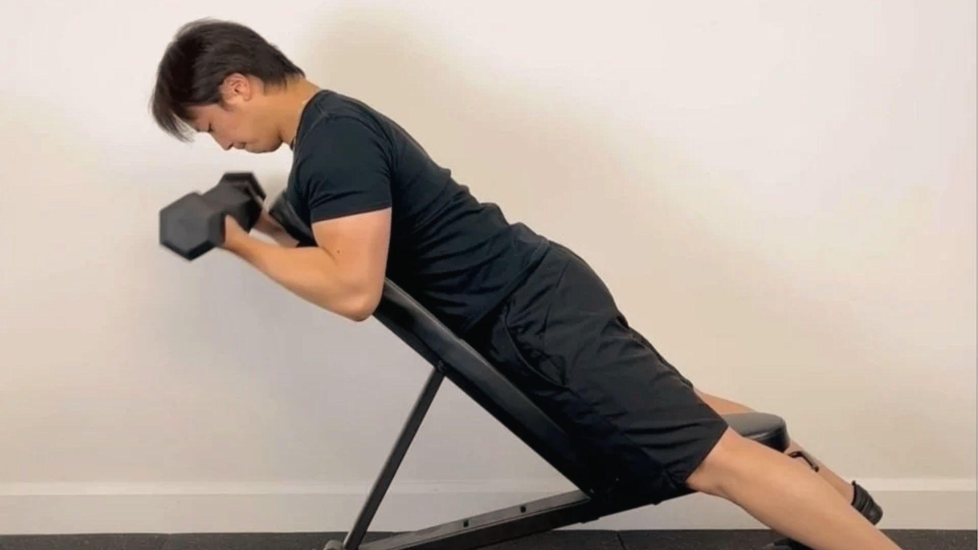 A man in black shirt performs dumbbell spider curls on an incline bench, chest pressed, arms curling forward.