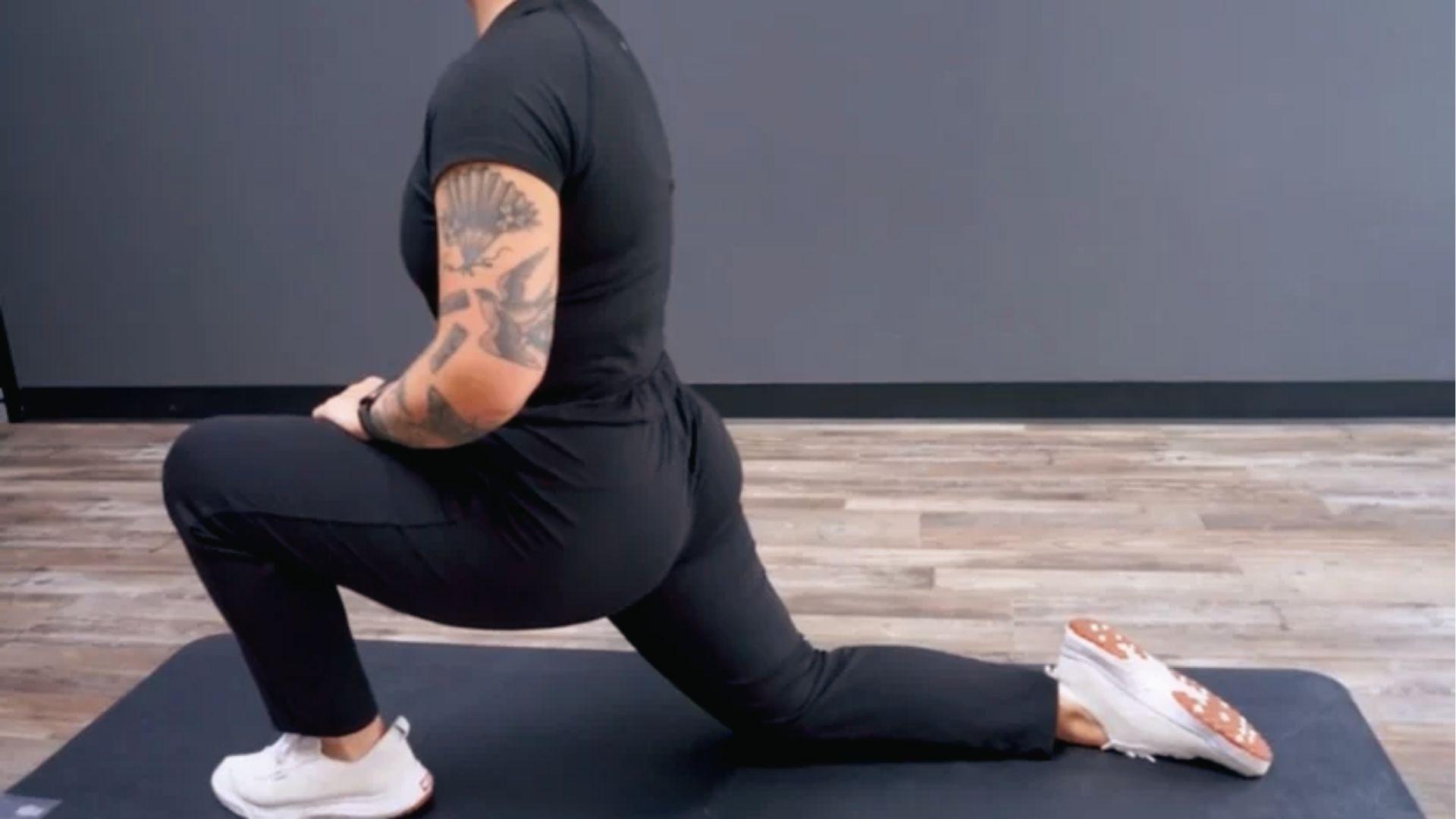 A person in black activewear does low lunge hip flexor stretch on the mat, front knee bent 90 degrees.