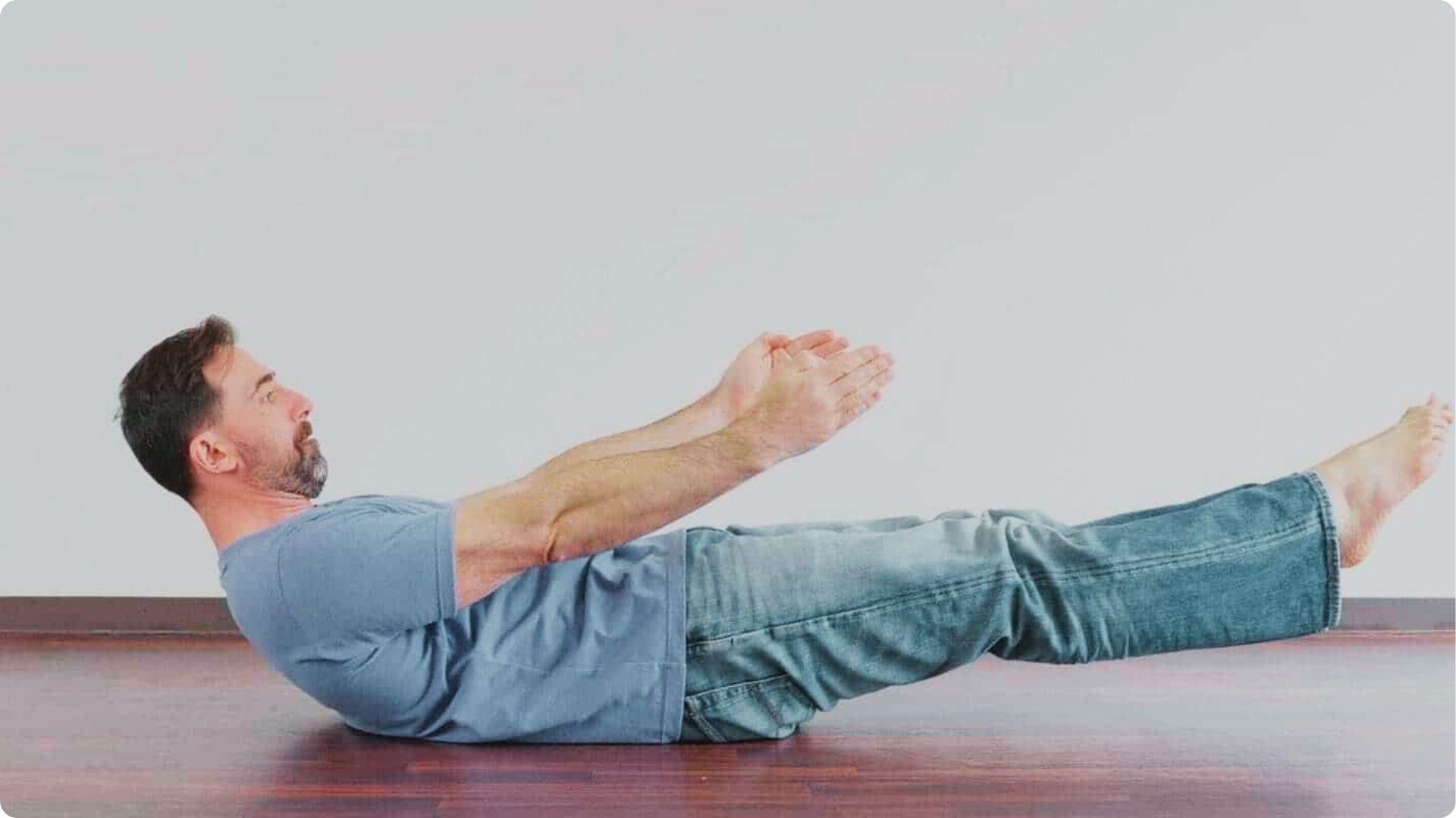 A man in a blue t-shirt and jeans demonstrates a hollow body hold on a hardwood floor.