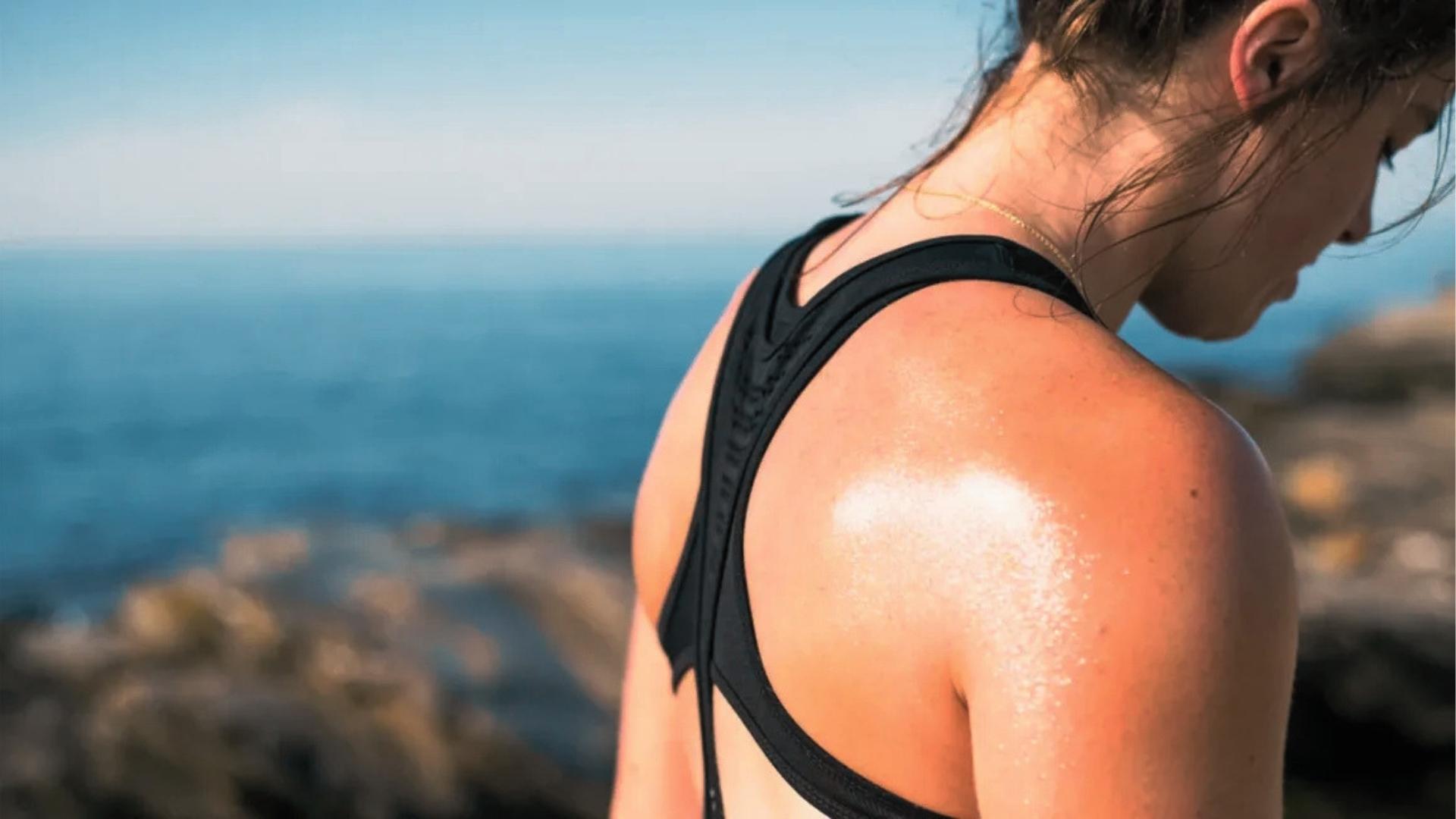 Woman in black sports bra stands by ocean cliffs with sweat glistening on sunlit shoulder after workout.