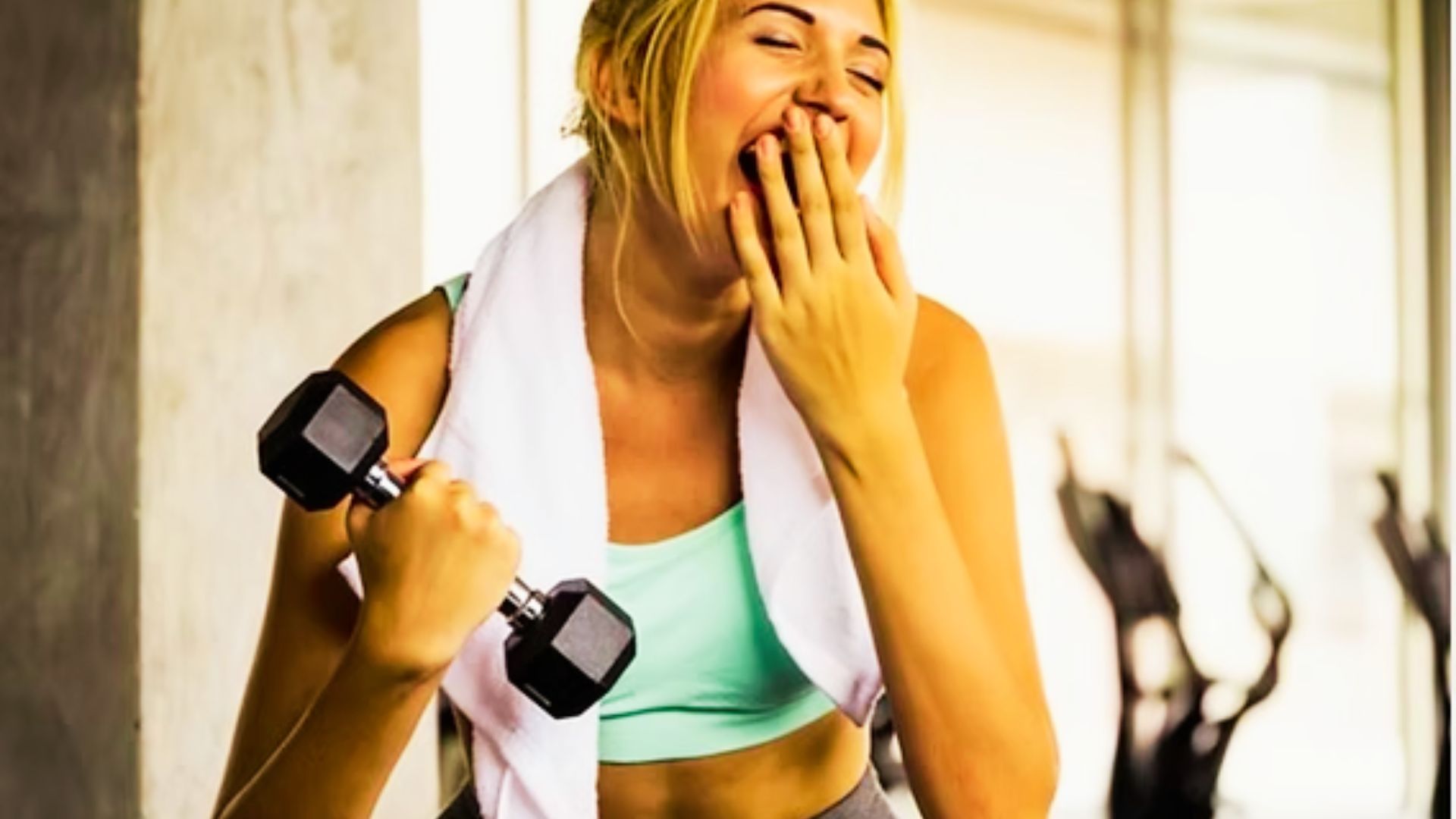 A woman in a teal sports bra holds a dumbbell and covers her mouth while yawning mid-workout in a gym.