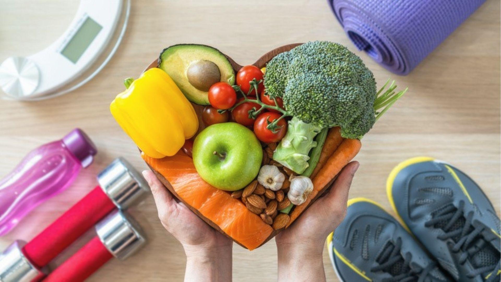 Hands hold heart-shaped bowl of veggies, salmon, nuts with dumbbells and shoes nearby for plan prep