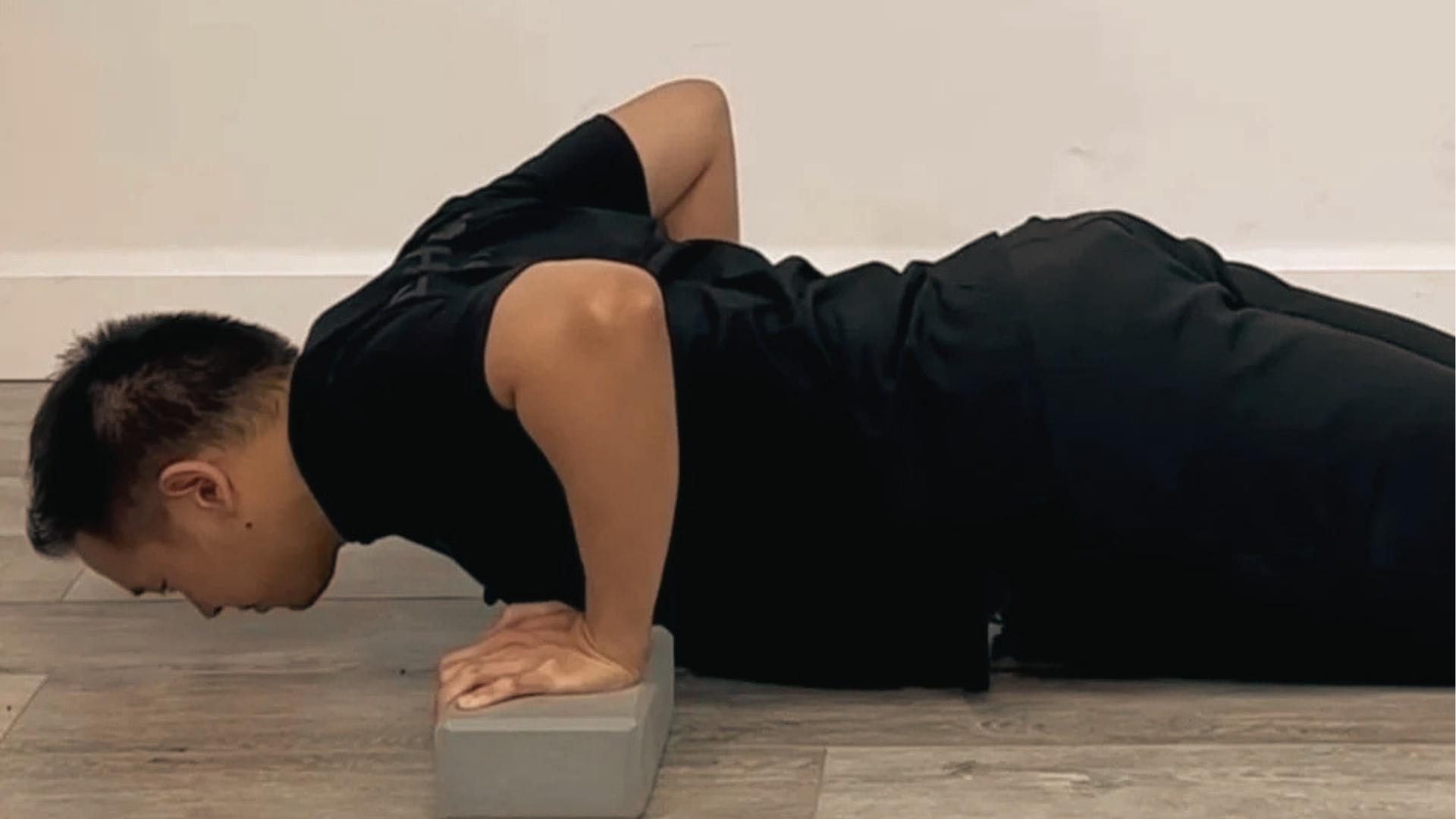 Man in black shirt performs deficit push-up variation using gray yoga block.