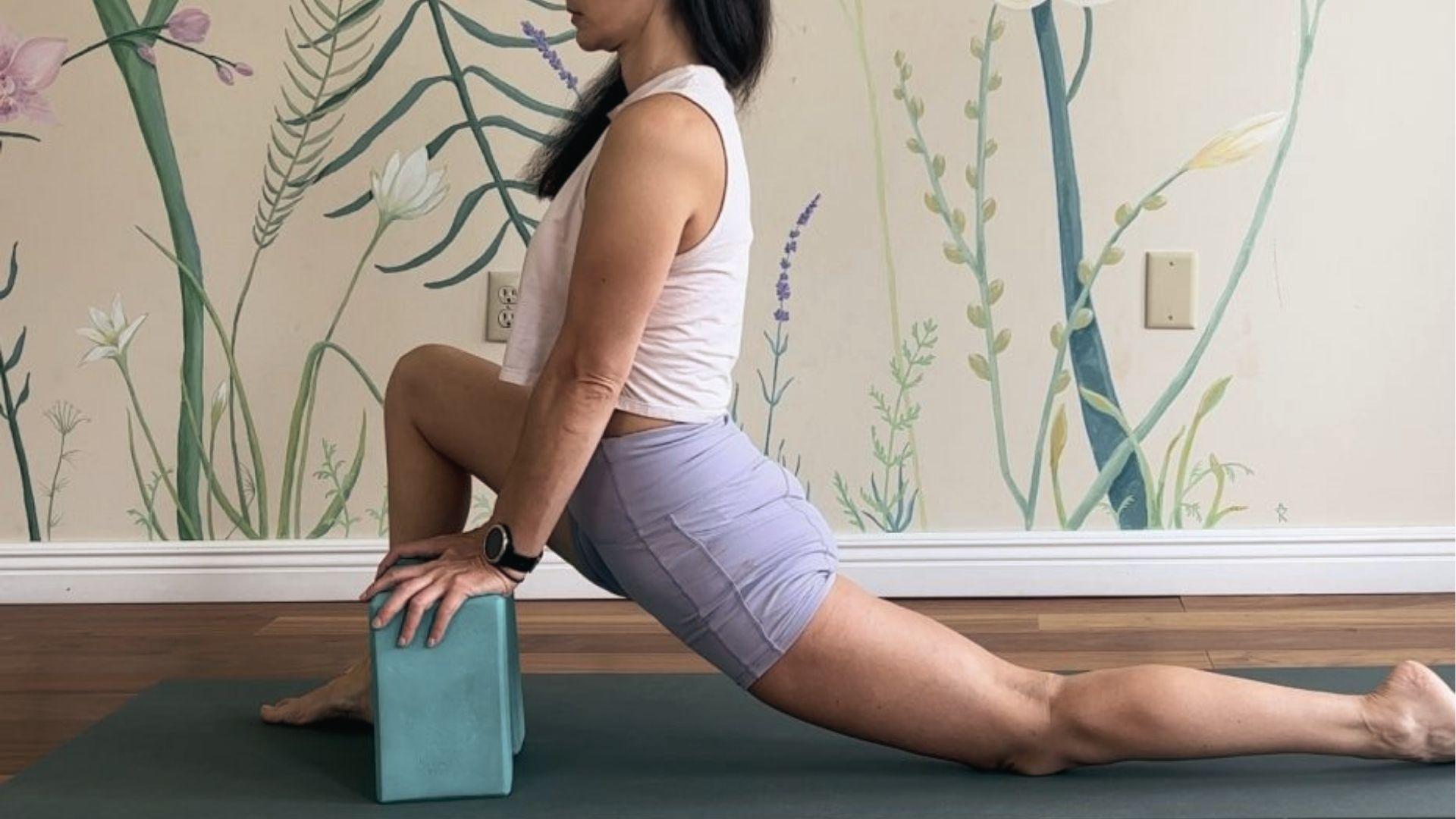 Woman uses yoga block for supported hip flexor lunge stretch on mat against floral wall mural.