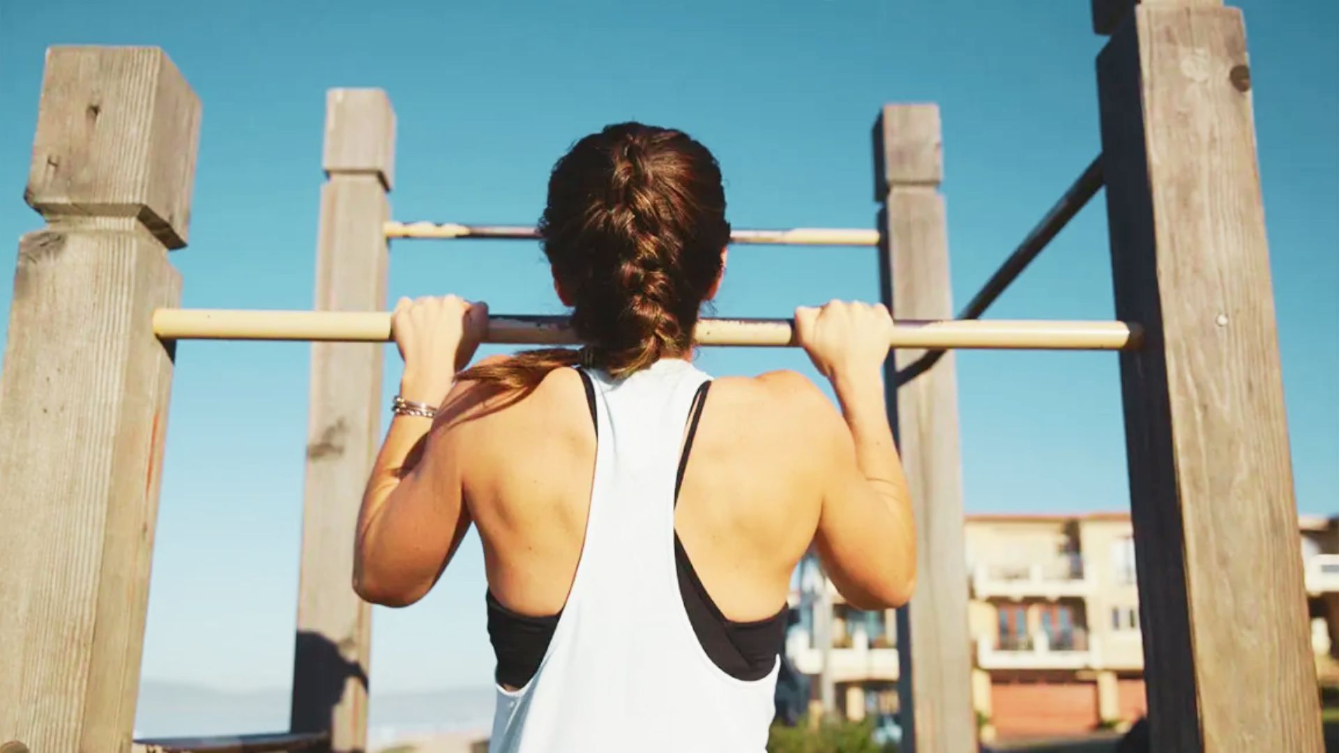 Muscular Woman doing wide-grip pull-ups on outdoor calisthenics bar under clear sky.