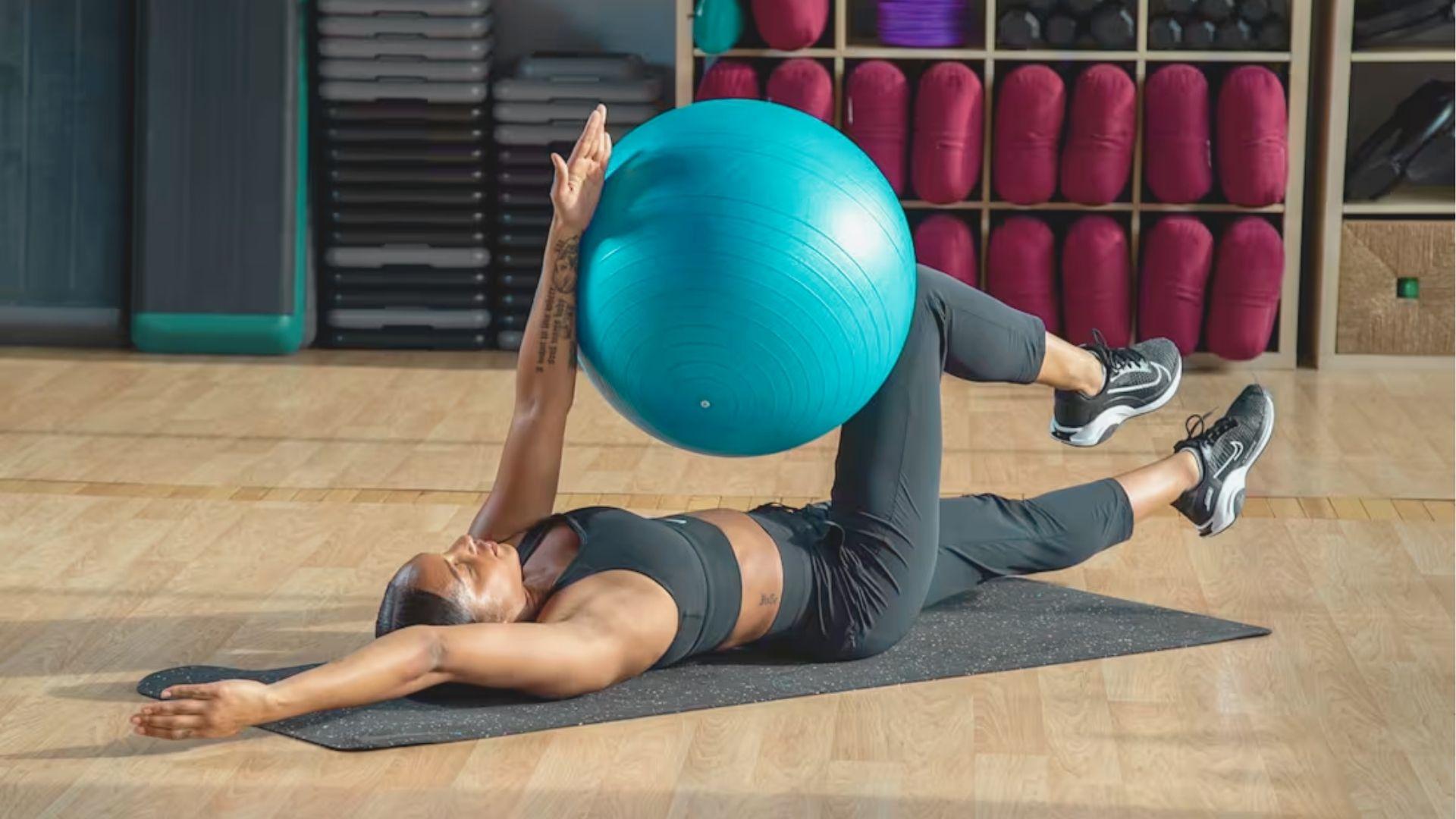 Woman in gym uses exercise ball for Dead Bug Exercise variation on yoga mat.