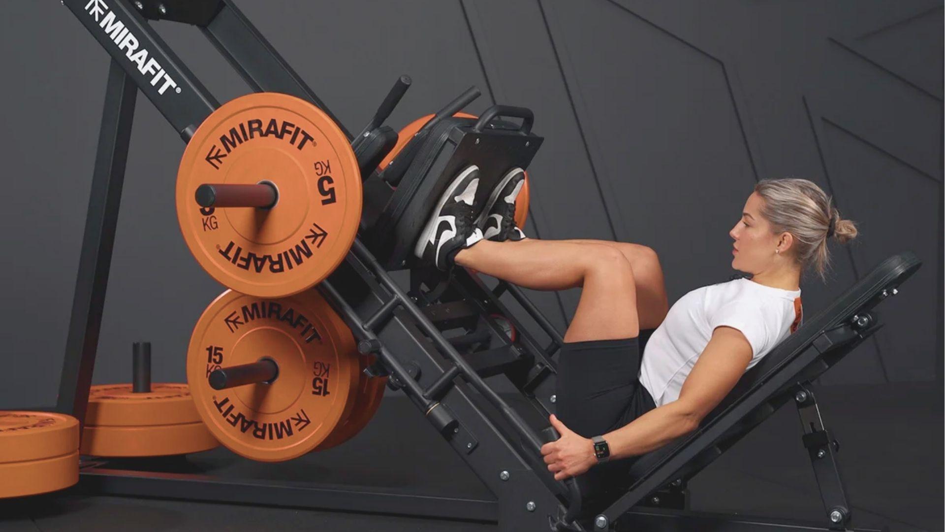 Woman using a leg press machine with orange weight plates in a gym.