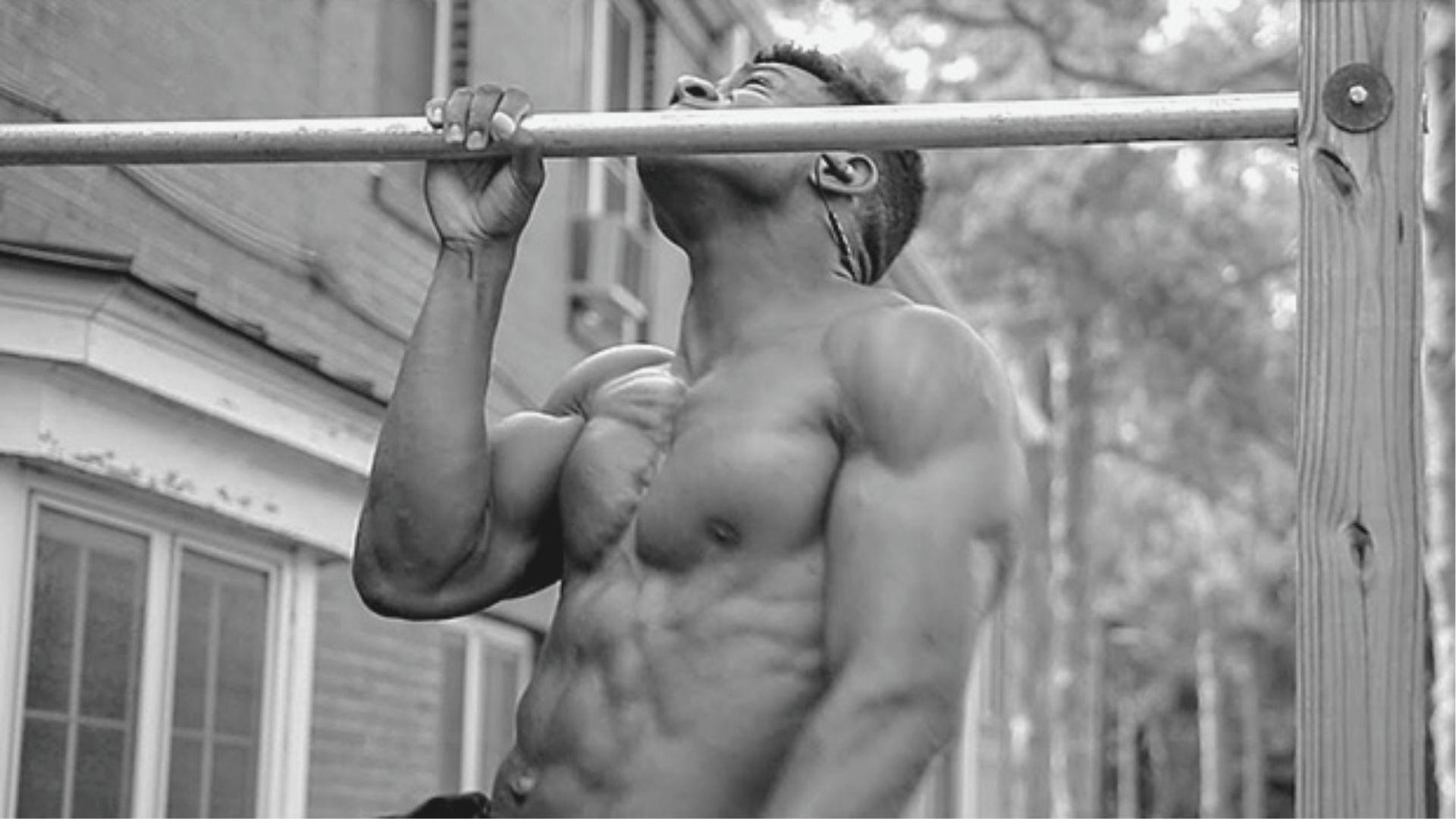 Black and white photo of shirtless muscular man completing a pull-up on an outdoor wooden bar.