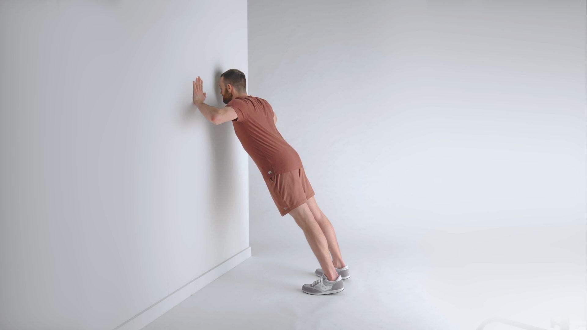 Man in brown workout set doing wall negative push ups against a white wall.