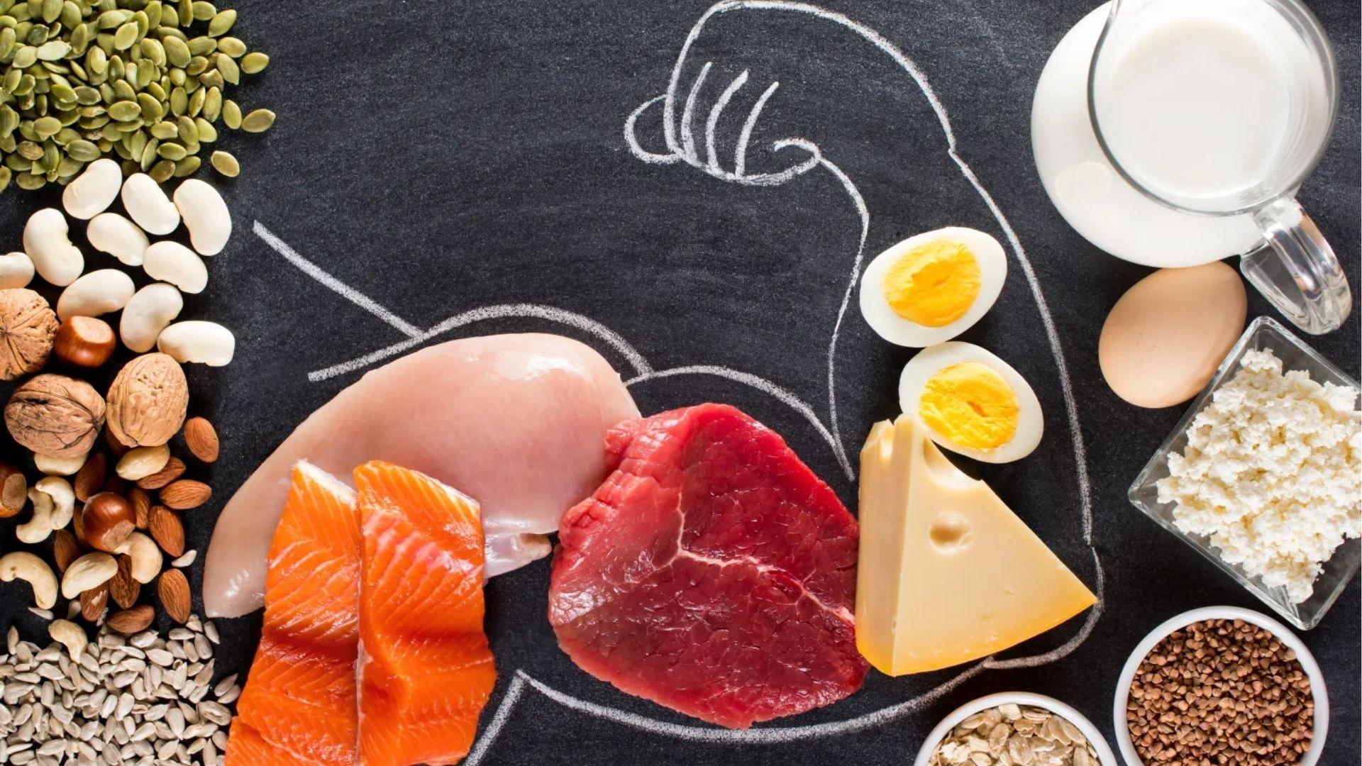 Salmon, chicken, beef, eggs, nuts, and seeds surround a chalk drawing of a flexed arm on a blackboard.