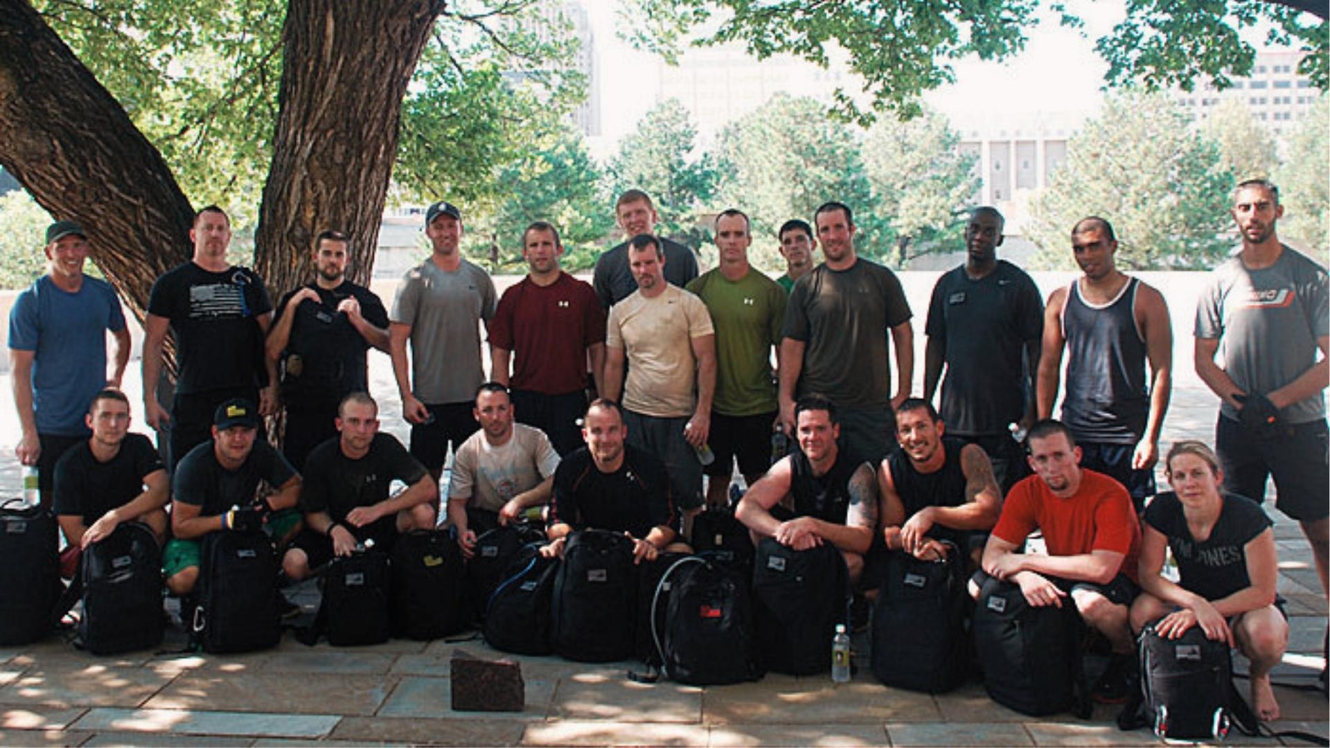 Group of men and women in athletic wear pose outdoors with weighted rucking backpacks for team fitness events.