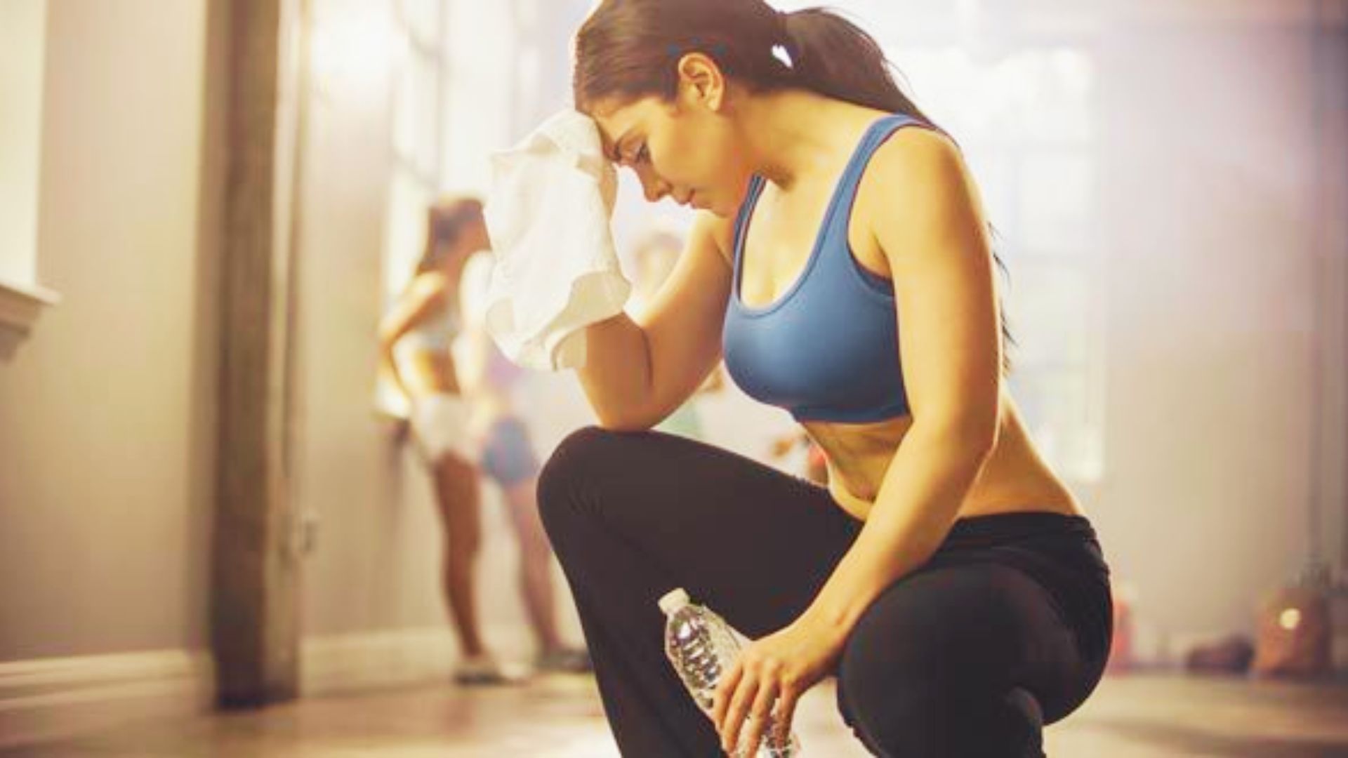 Woman in a blue sports bra wipes sweat from her forehead while holding a water bottle, looking fatigued.