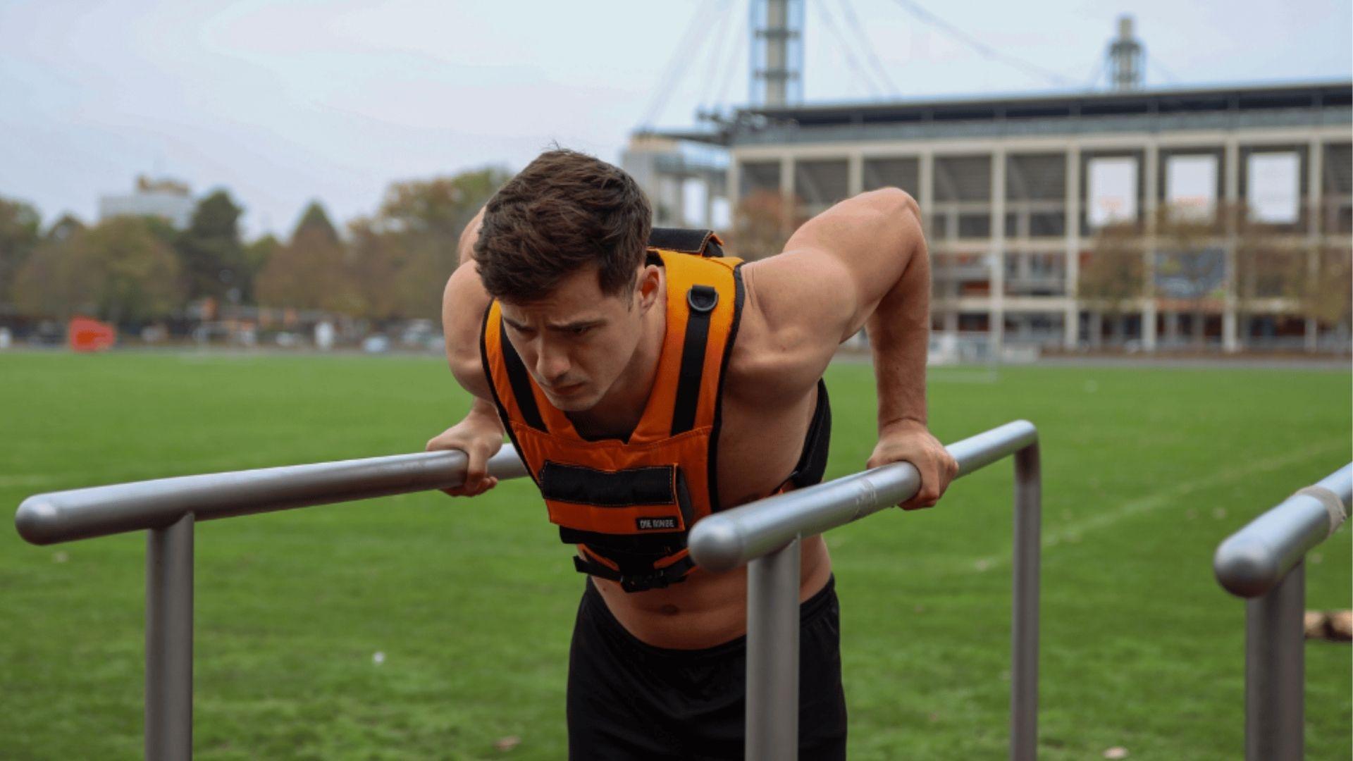 Man in an orange weighted vest performing a negative dip on parallel bars outdoors at a stadium.