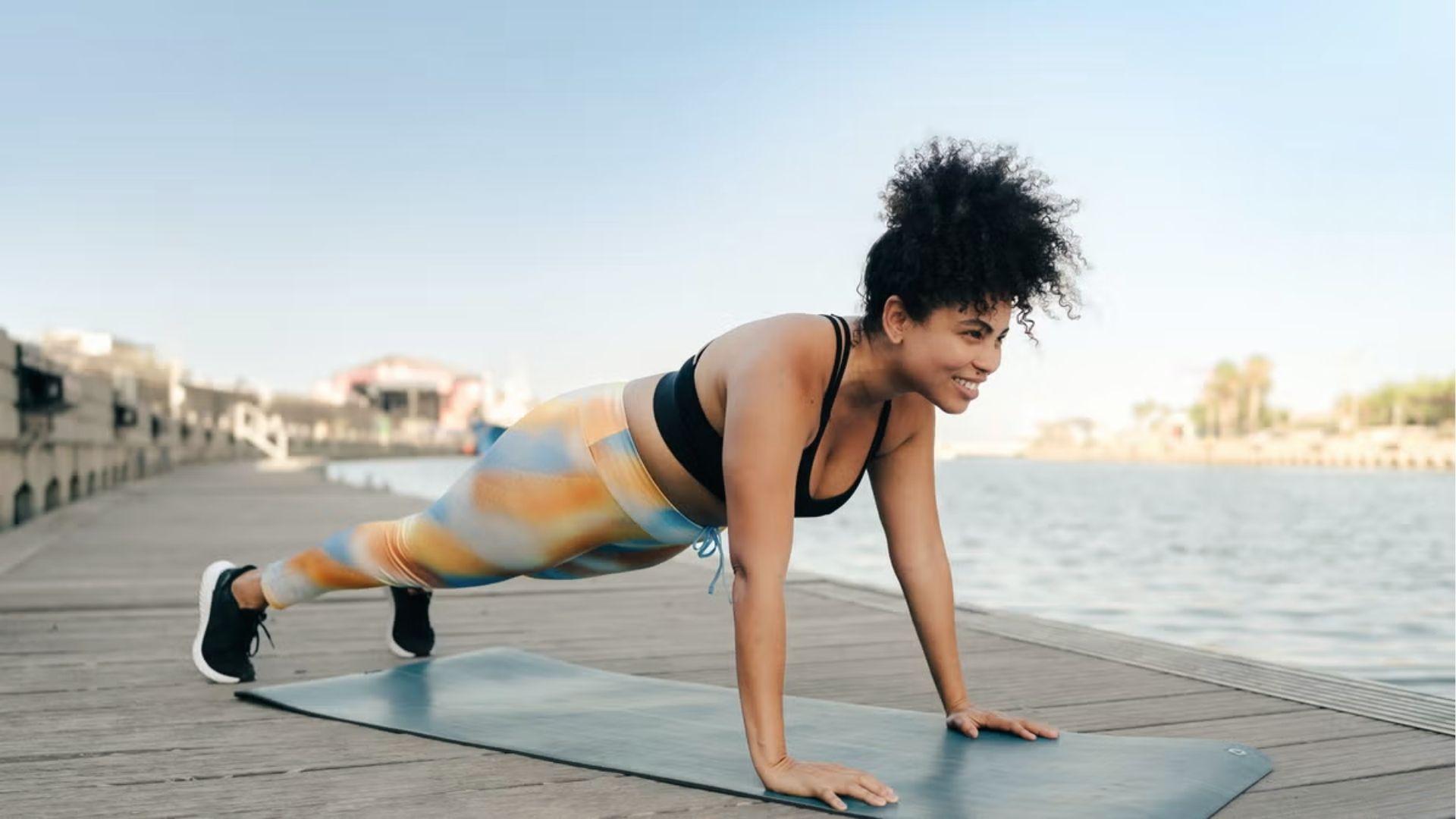 Woman holds high plank on mat by water for core stability day in beginner calisthenics routine