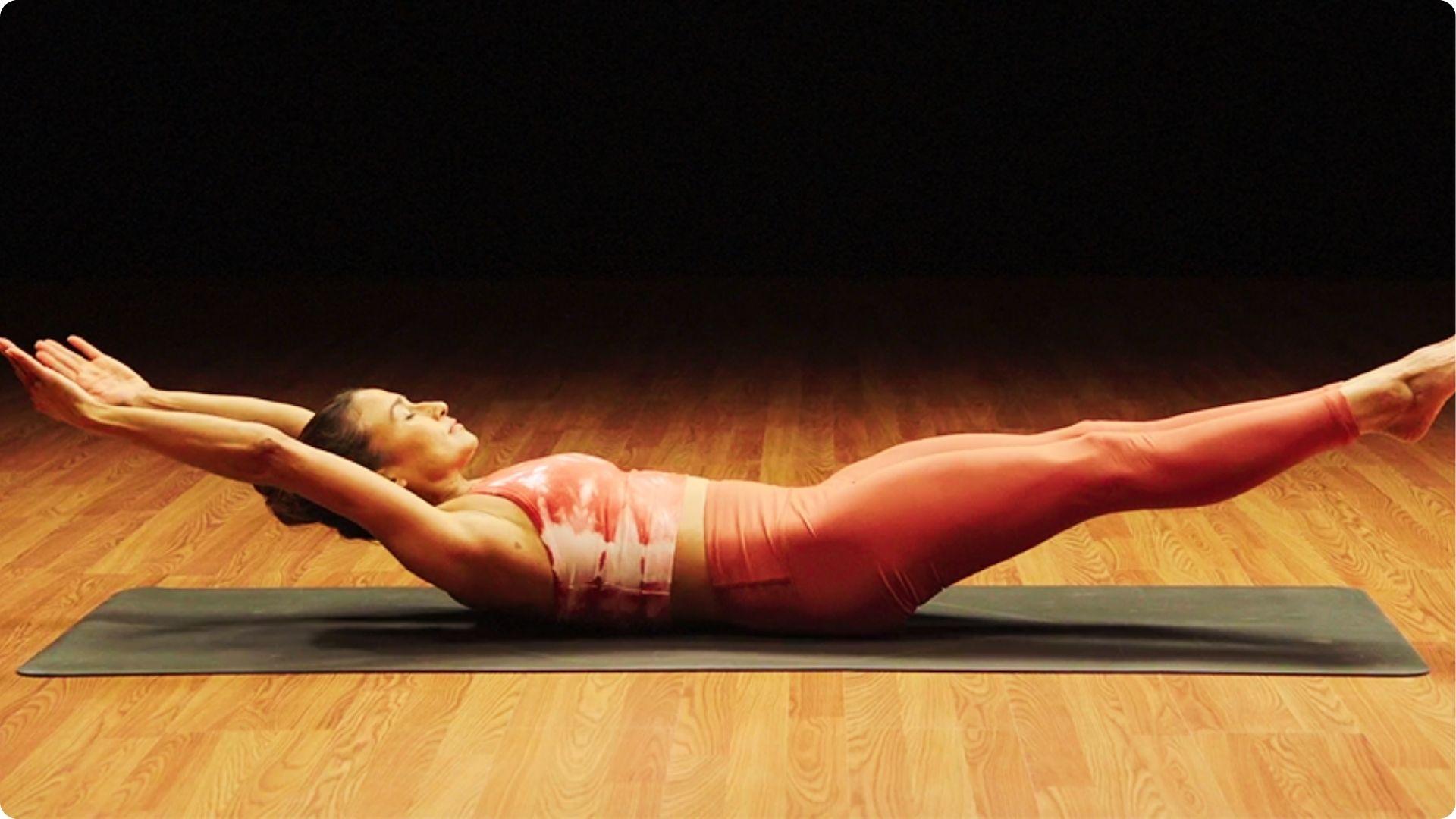 A woman in a coral tie-dye top and matching leggings holds a full hollow body position on a mat.