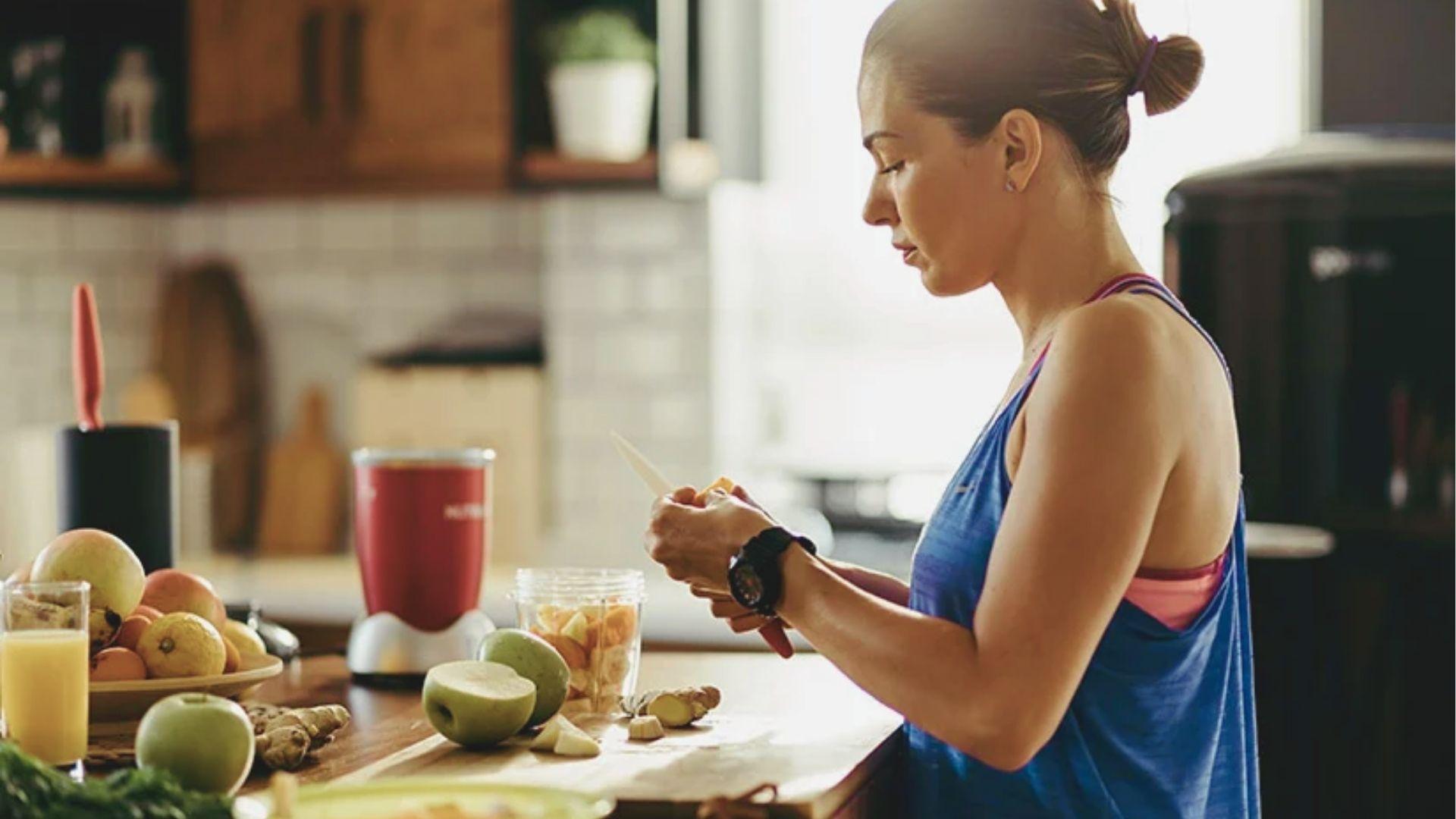 Woman in blue tank top prepares a fruit smoothie in a sunlit kitchen with fresh apples and ginger nearby.