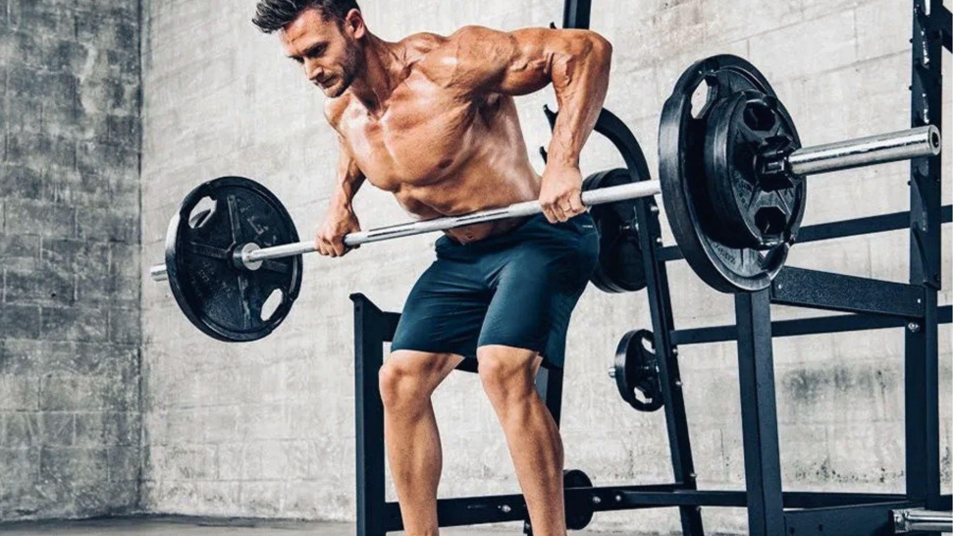 Shirtless man performs bent over barbell row with overhand grip in industrial gym near rack