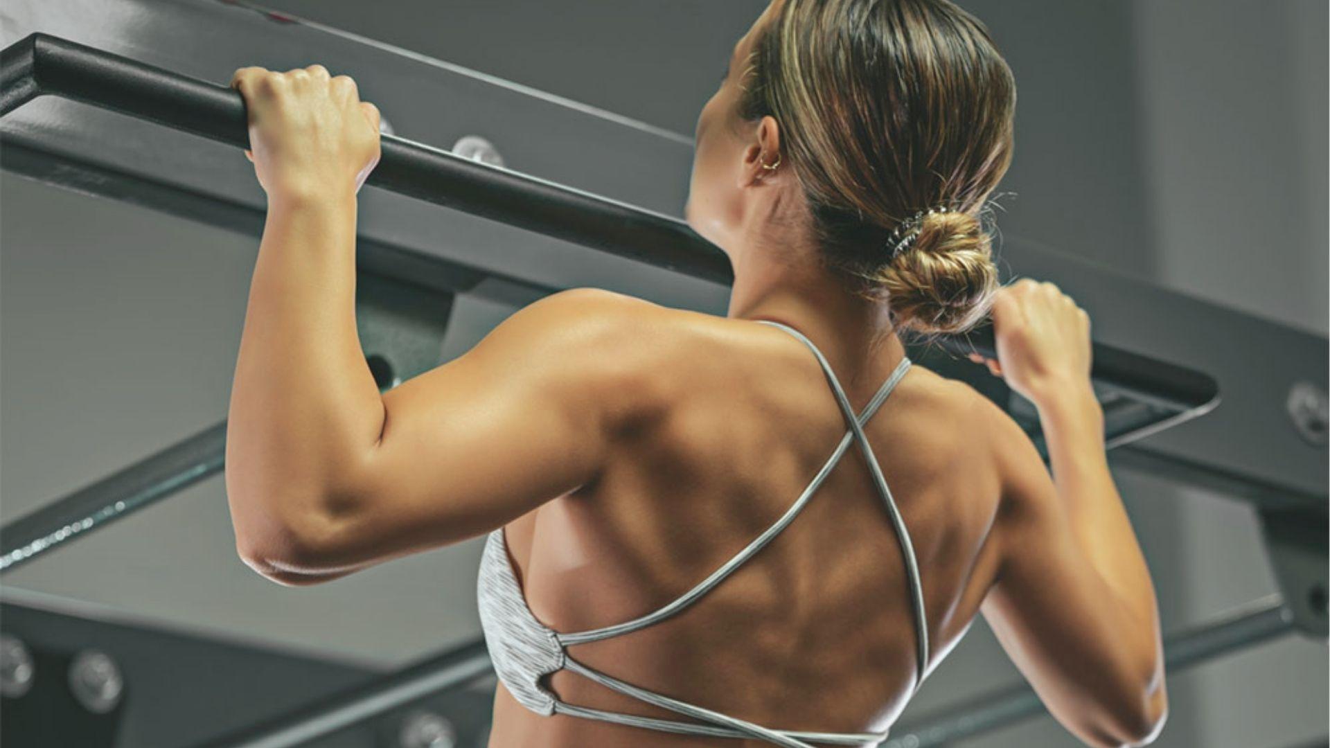 Woman in a white strappy sports bra shown from behind, pulling herself up on a pull-up bar.