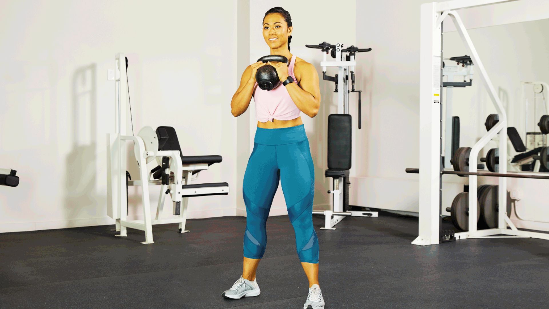 Woman standing in a gym holding a kettlebell in the goblet position at chest height, preparing for a squat.
