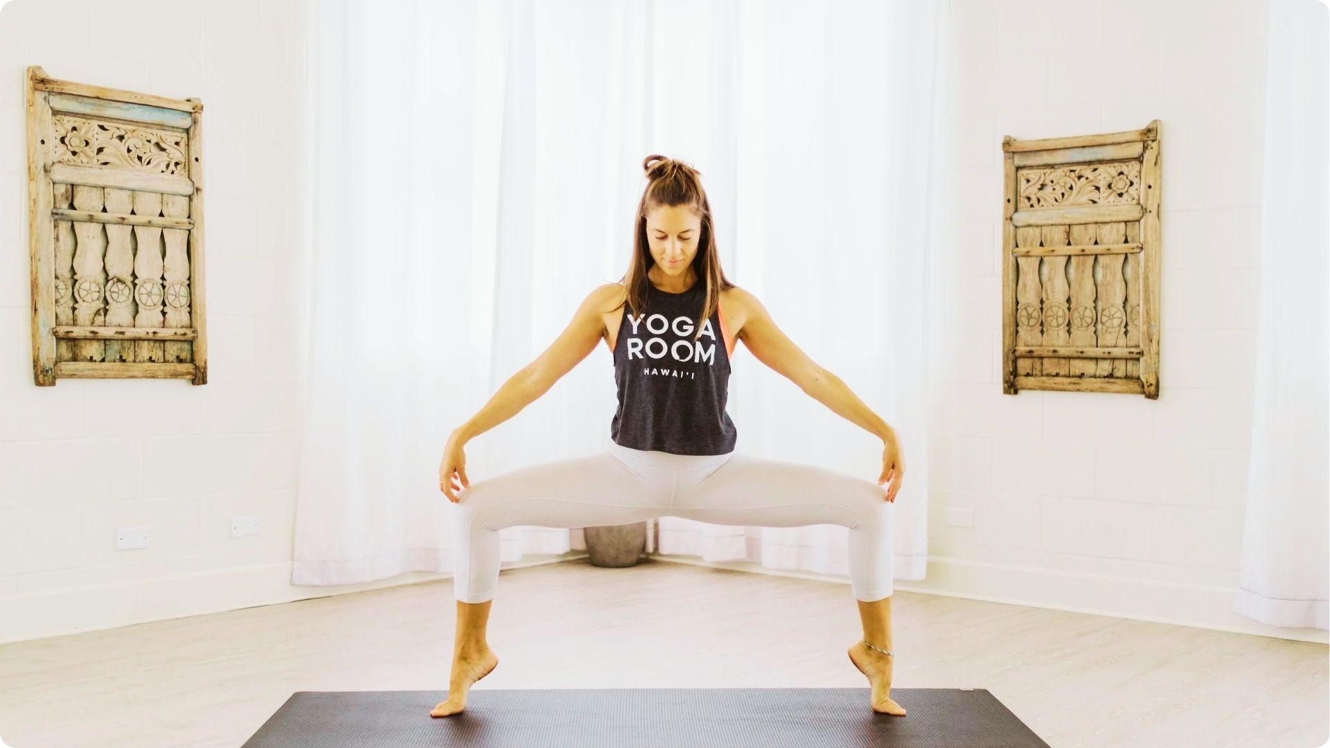 Woman in a "YOGA ROOM HAWAI'I" tank top and white leggings holding Goddess Pose on a mat with heels lifted.