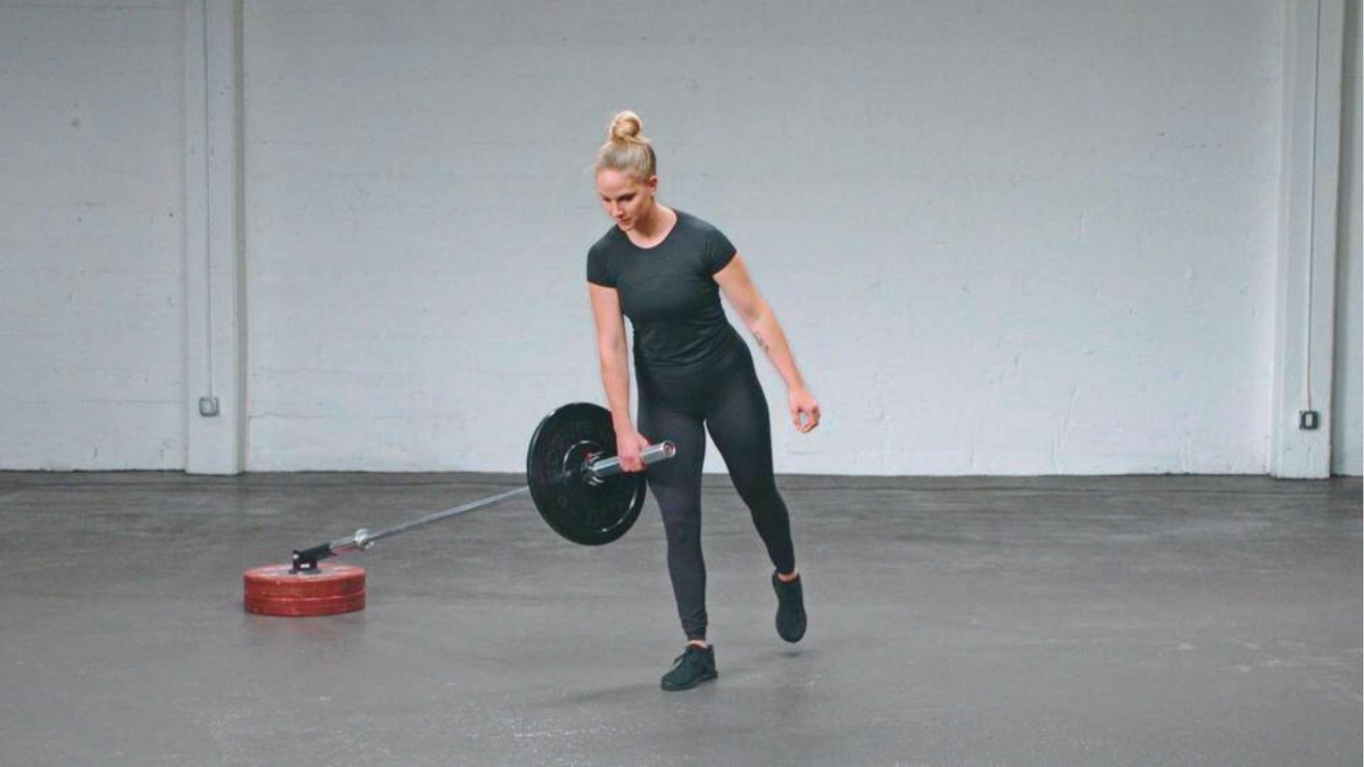 A woman in black workout clothes balances on one leg while holding a landmine barbell in one hand.