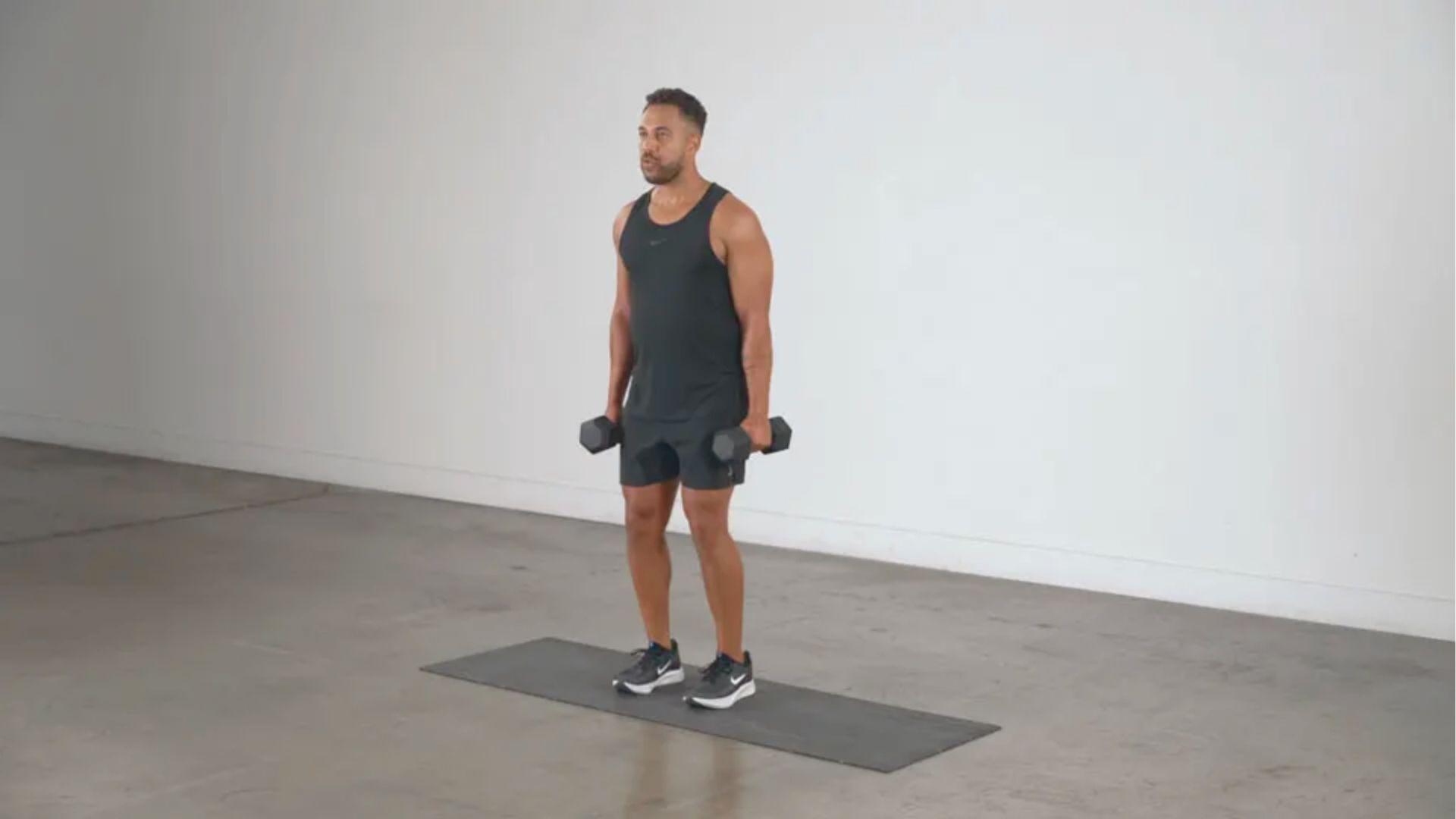 Man in black tank top holds dumbbells while standing on an exercise mat in a studio.