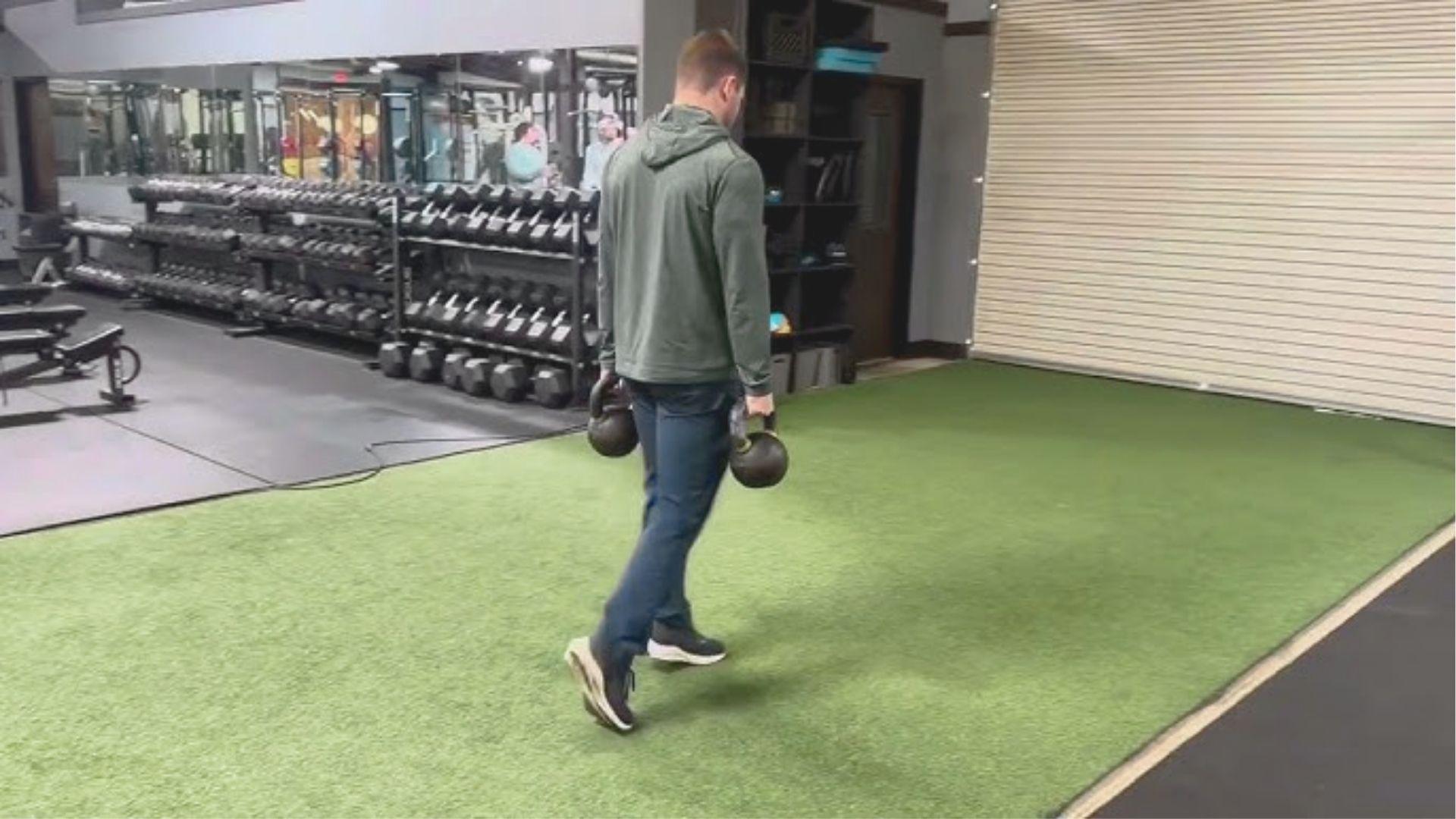 Man in a green hoodie walks across gym turf while holding two kettlebells at his sides.