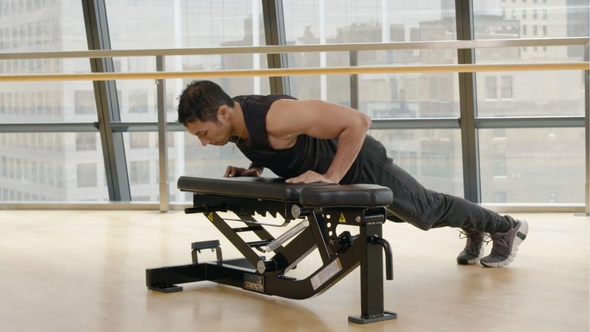Man in black tank top performs incline push up on flat gym bench near large windows with city view