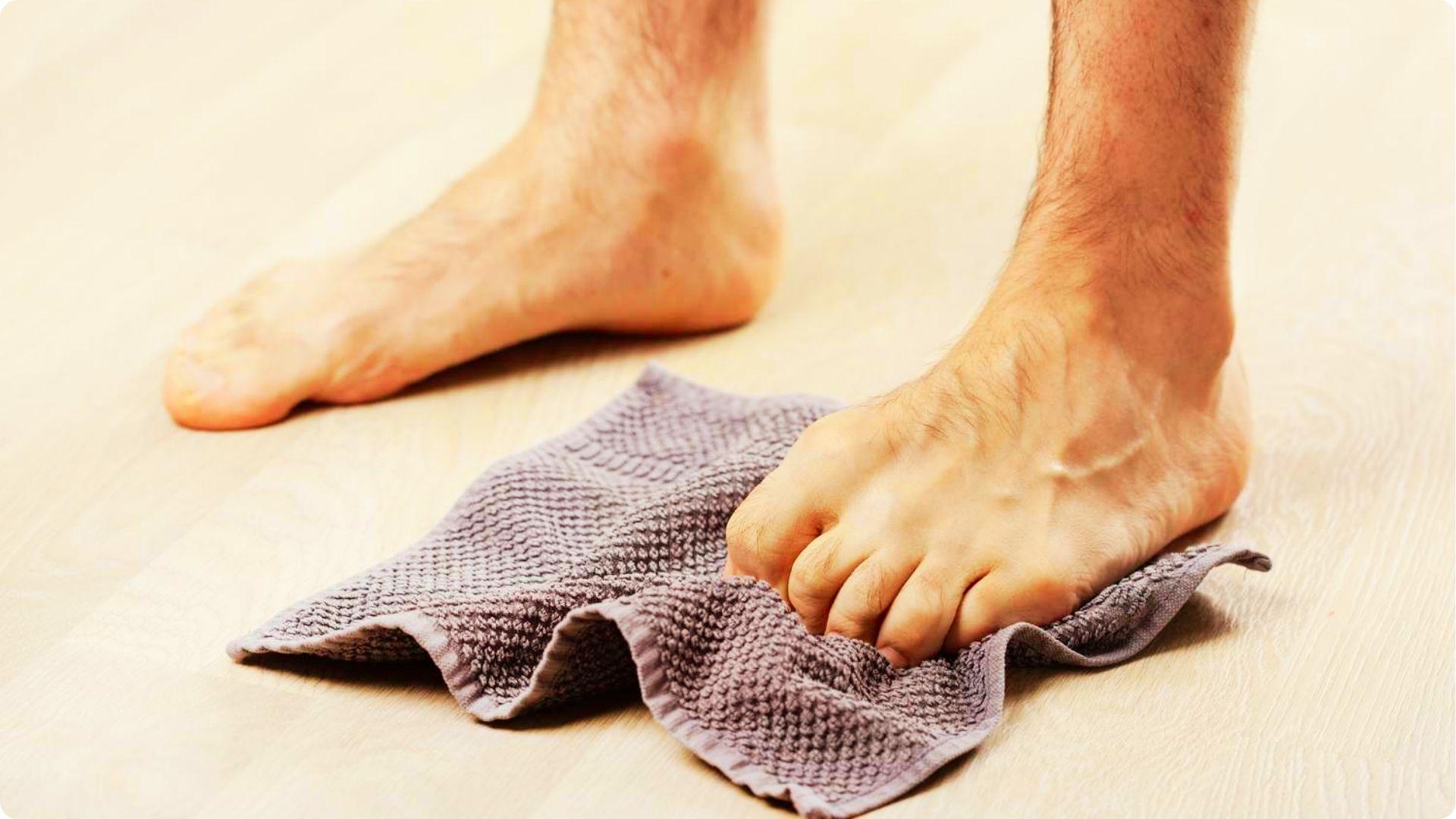 Person uses bare foot to scrunch a grey towel on a wooden floor with toes curled.