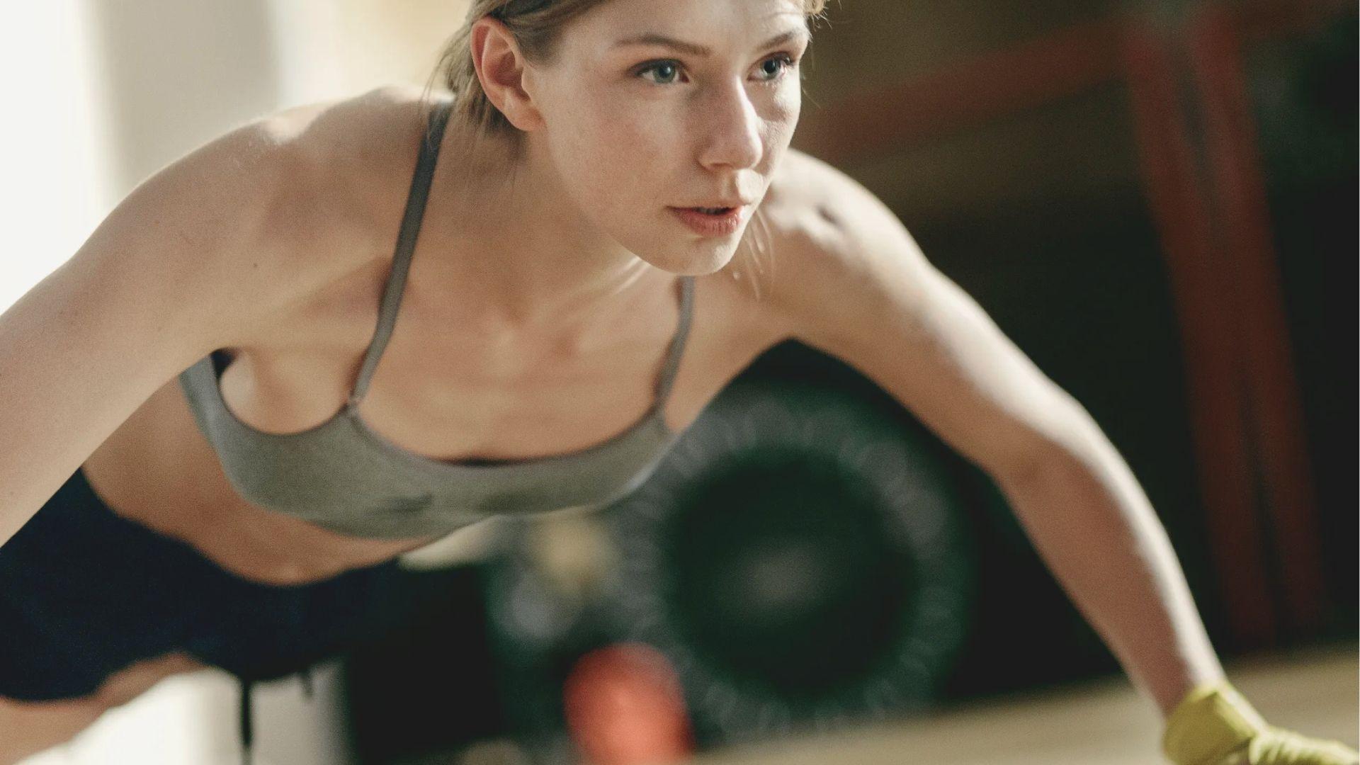 Woman in a gray sports bra performing a wide-arm push-up variation indoors.