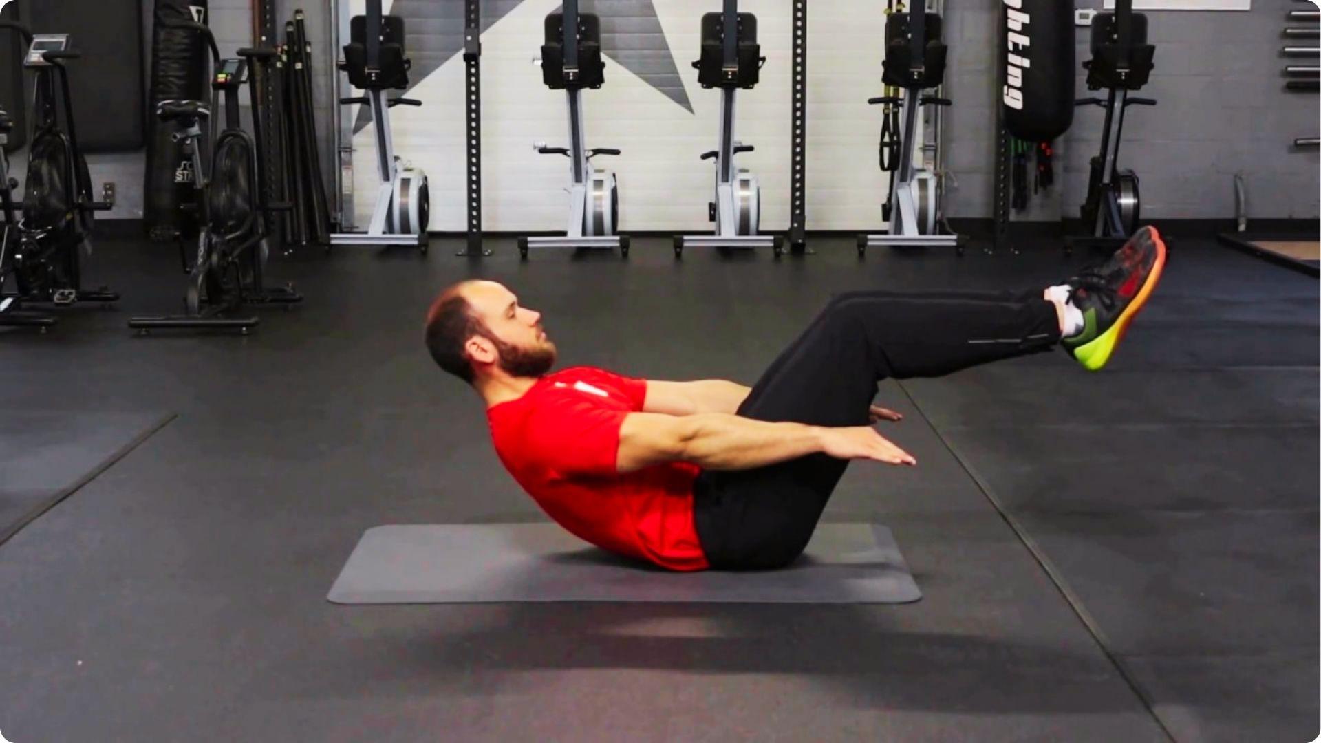 A man in a red shirt and black pants holds a bent-knee hollow body hold on a gray mat in a fitness studio.