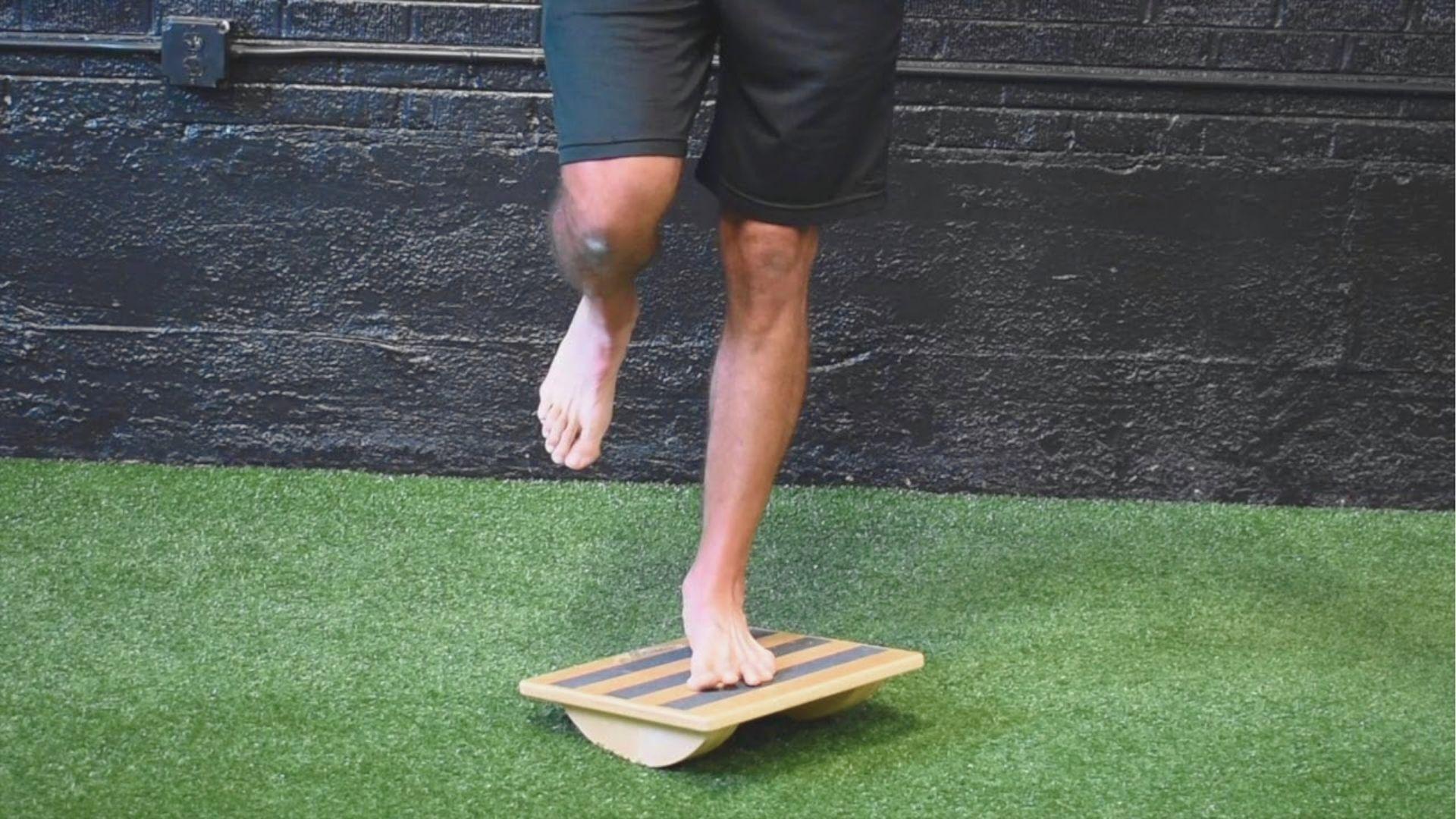 Man balancing barefoot on one foot atop a wooden wobble board with black stripes on artificial turf.