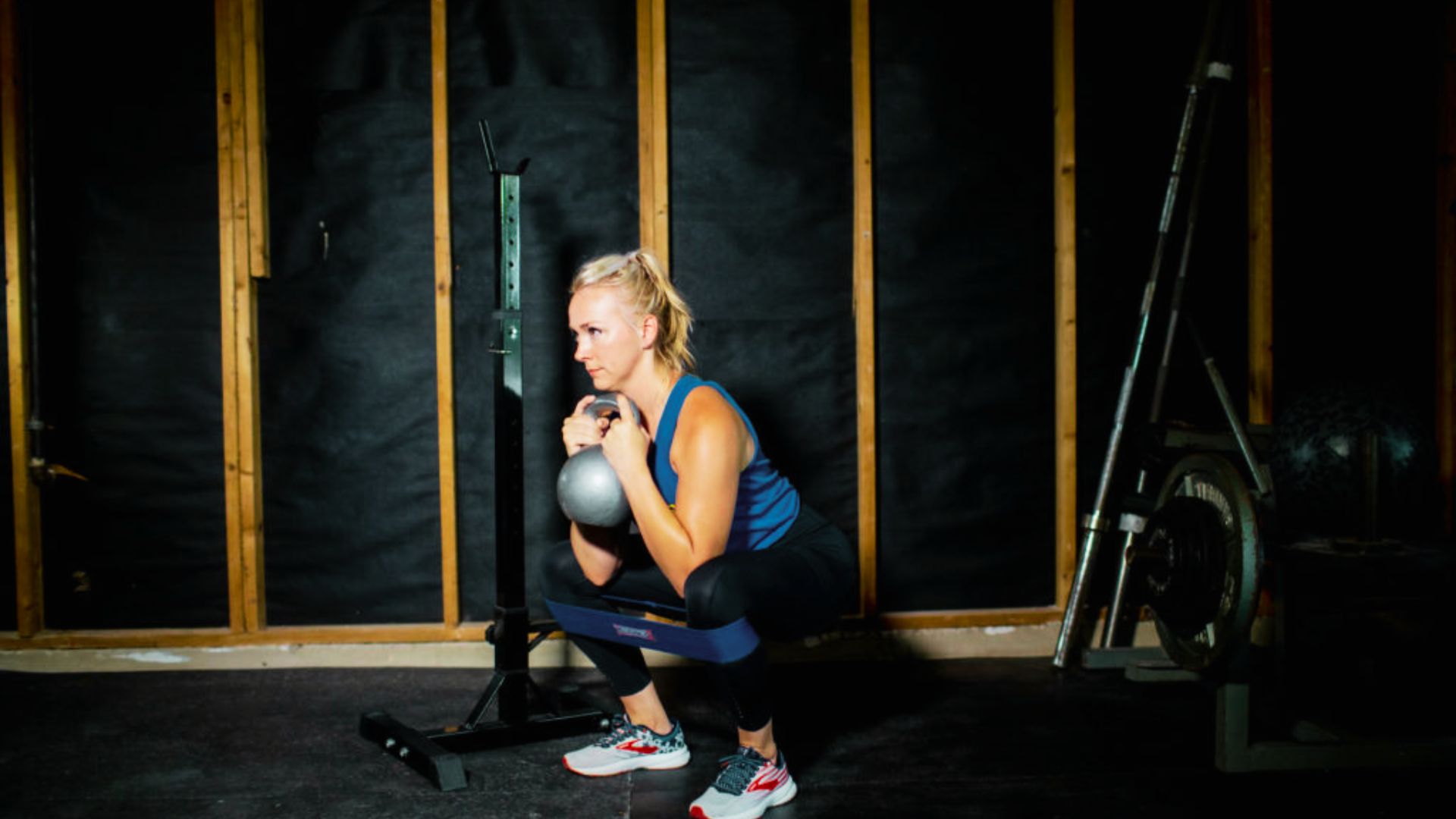 Woman performing a kettlebell goblet squat in a home gym, holding the weight at chest level with a resistance band around her thighs.