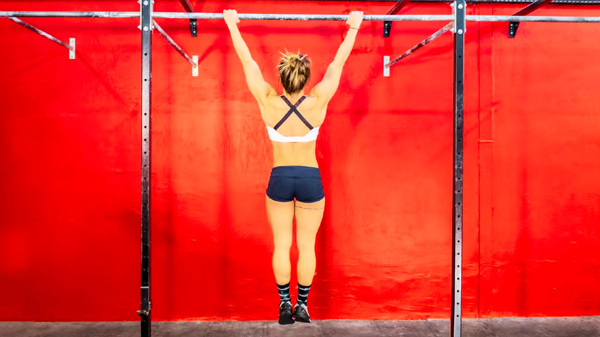 A woman hangs from a pull-up bar with arms fully extended, facing a bright red gym wall.