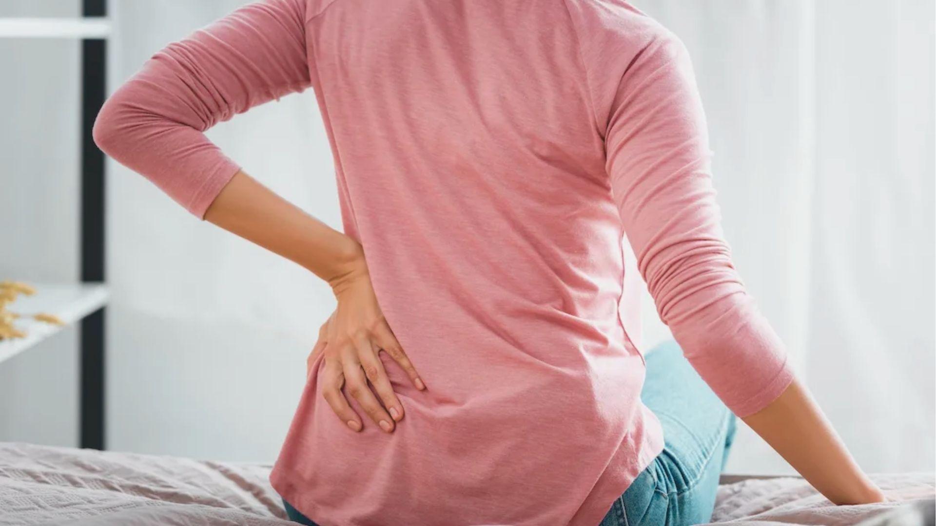 Woman in pink top sits on a bed, hands on lower back, stretching to relieve discomfort.