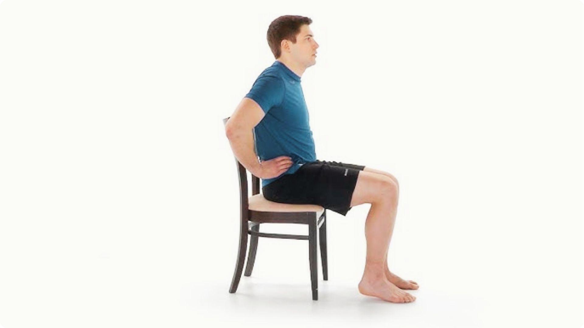 A man sits upright on a chair with hands on his lower back and feet flat on the floor.