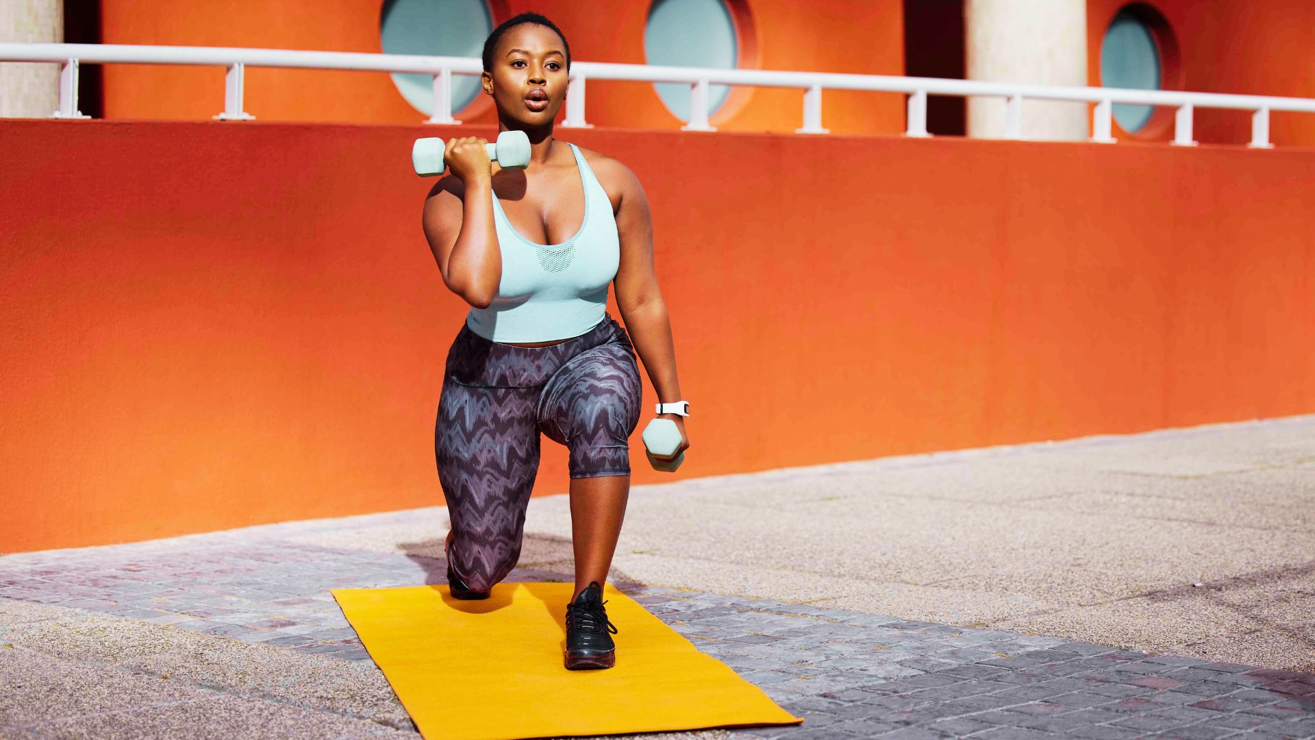 Strong woman in teal sports bra performs a weighted lunge on a yellow mat outdoors.