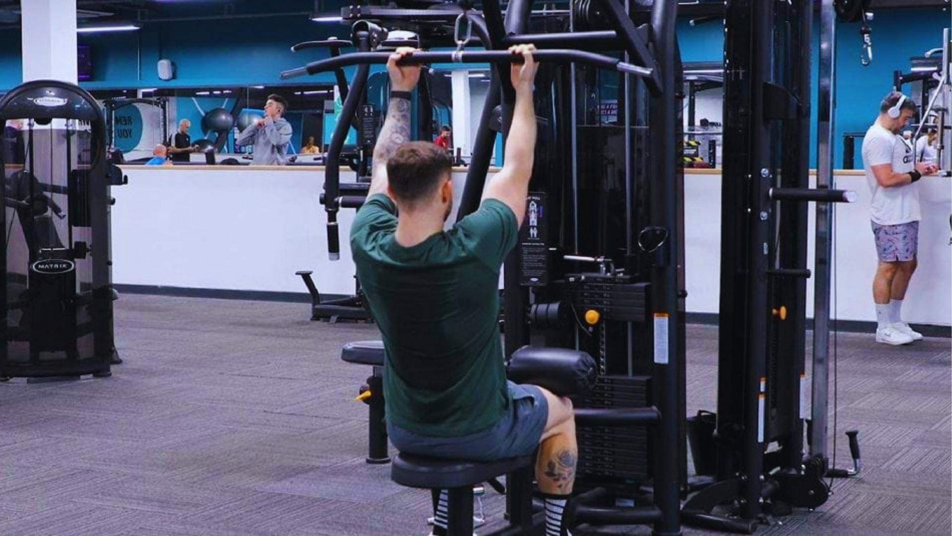 Man in green shirt uses overhand grip on lat pulldown machine with knee pads in busy commercial gym setting.