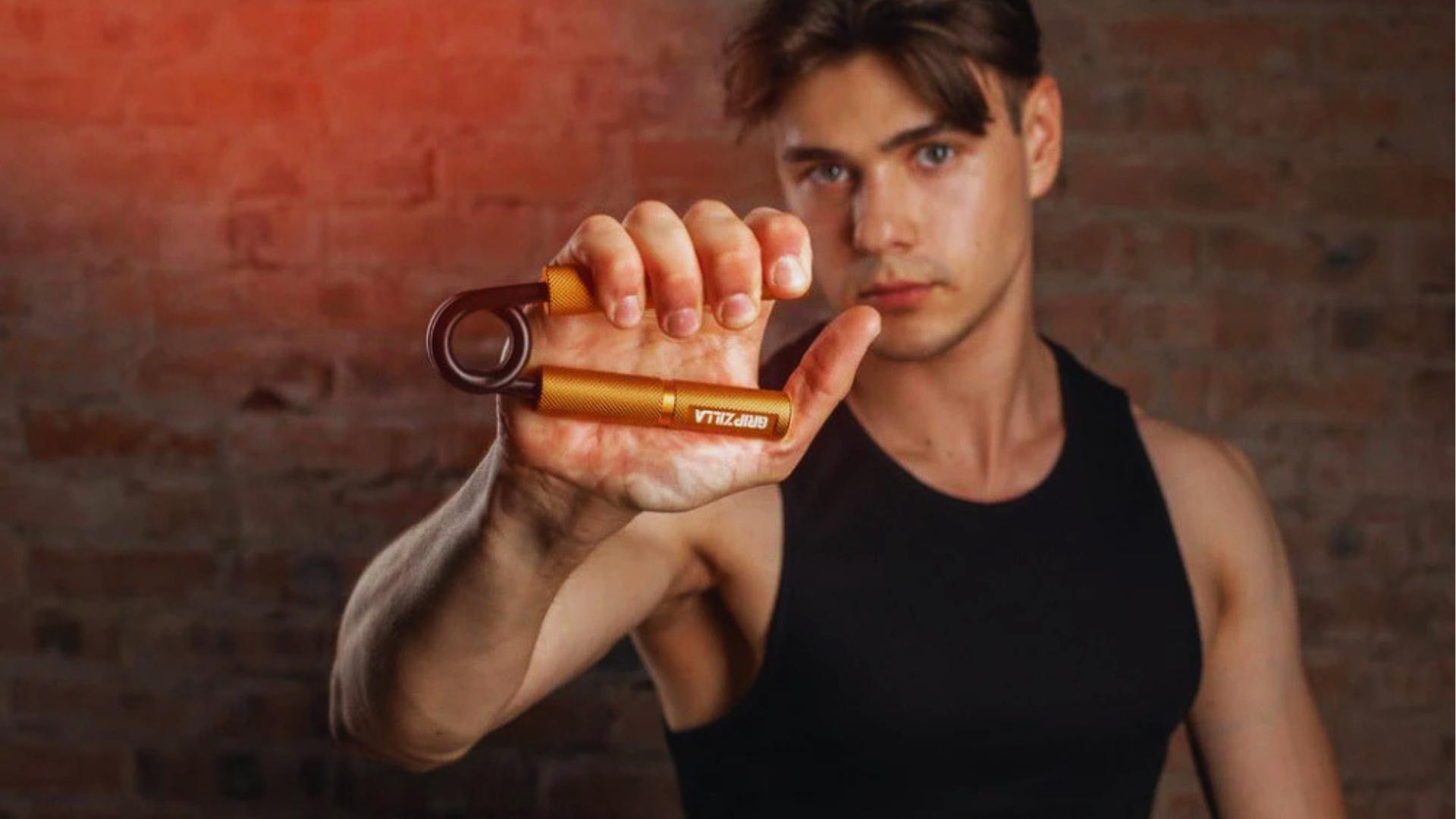 Young man in black tank top holds a gold Gripzilla hand gripper toward camera in front of exposed brick wall.
