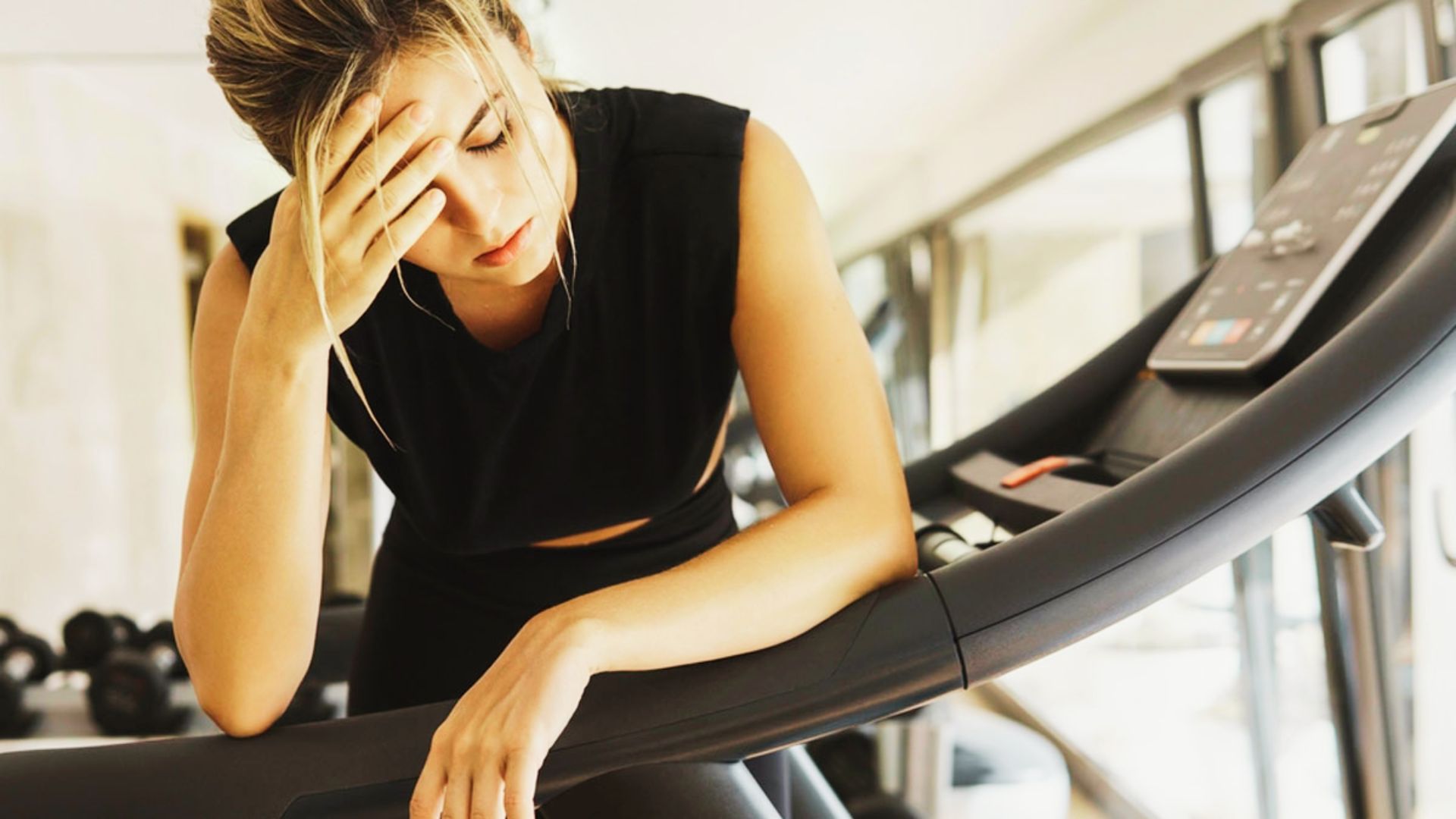 Woman in black workout clothes leans on a treadmill, holding her head as if dizzy or drained after cardio.