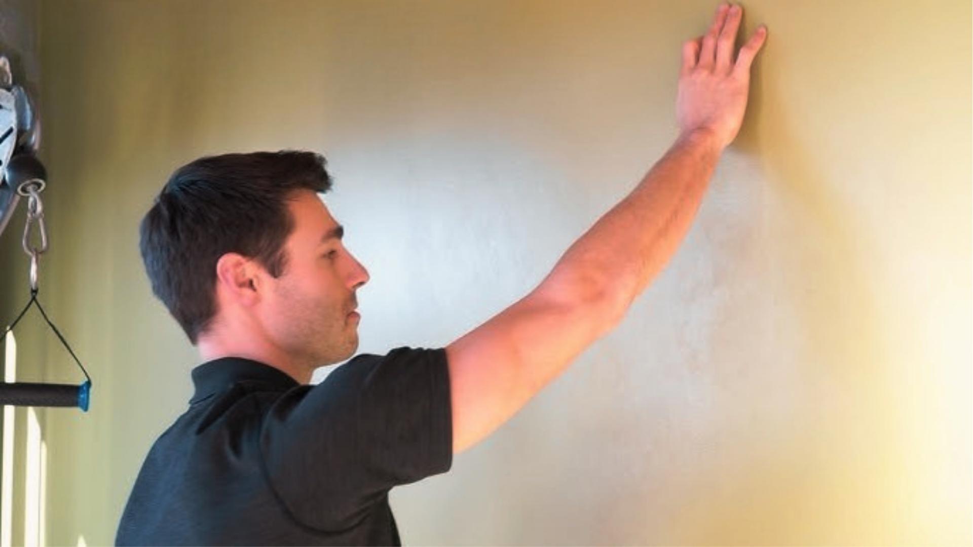 Man in black shirt does shoulder wall walk exercise, gradually sliding hand up wall to improve mobility.