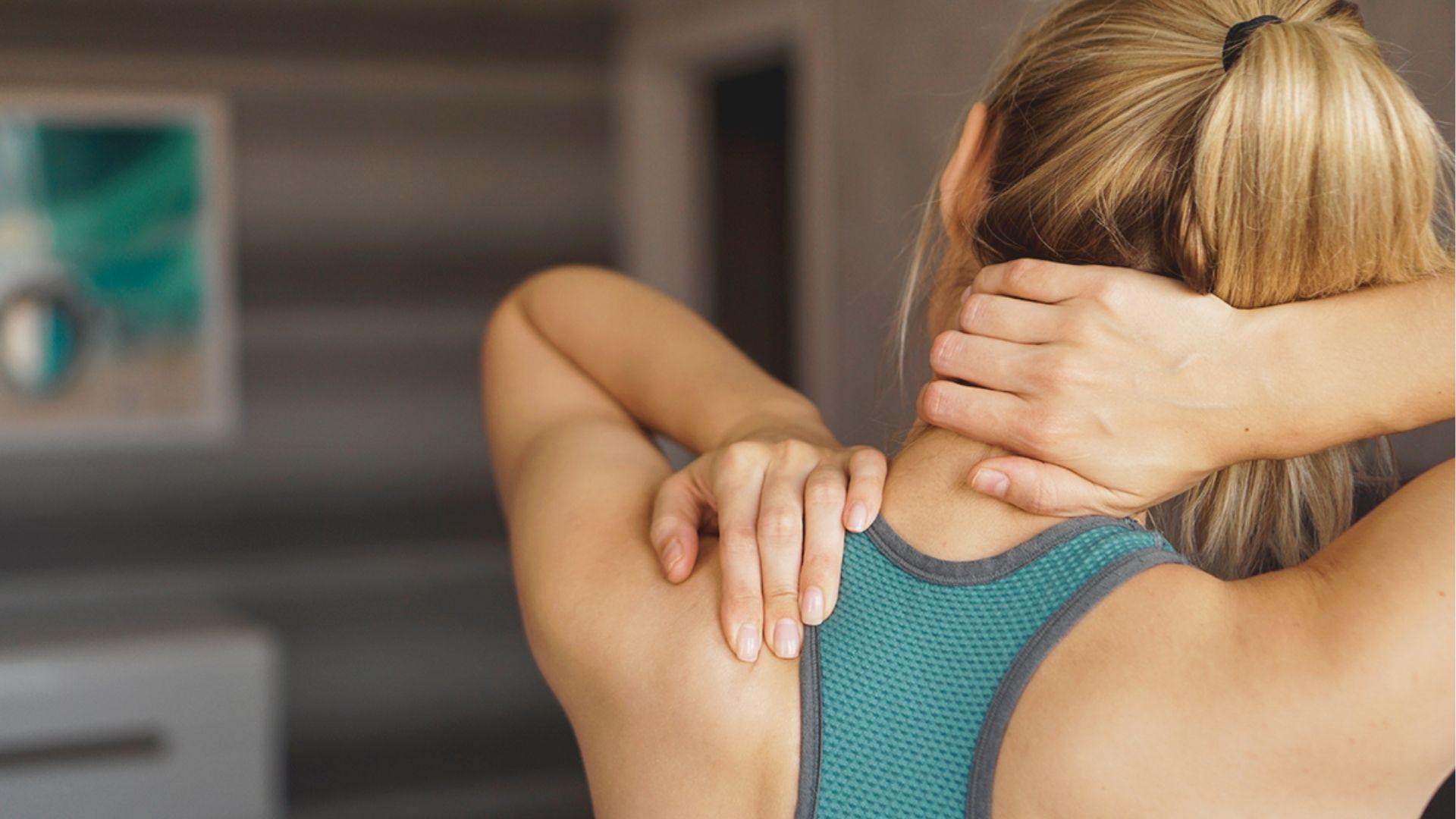 Woman in a teal sports bra grasps the back of her neck and upper shoulder with both hands.