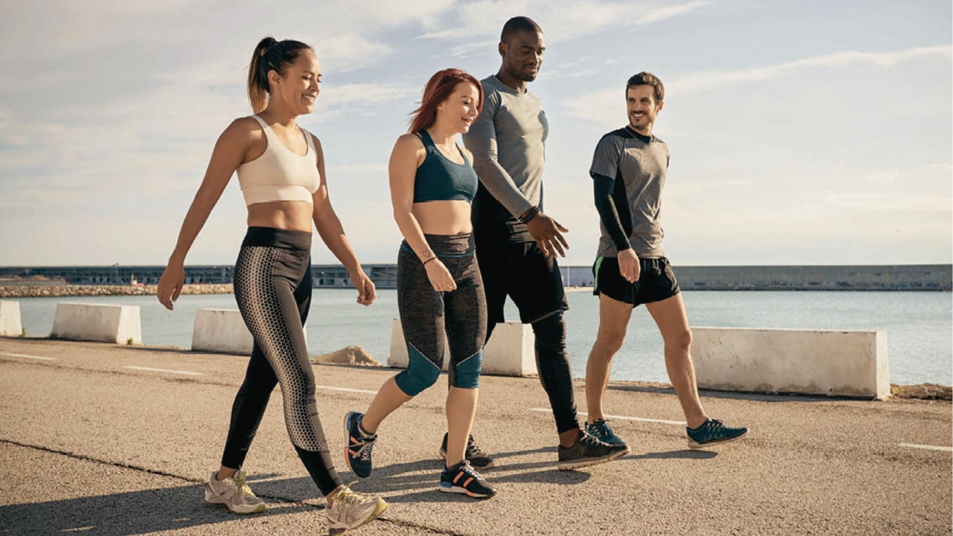 Diverse group of four friends in activewear power walks and chats along sunny waterfront promenade with city backdrop.