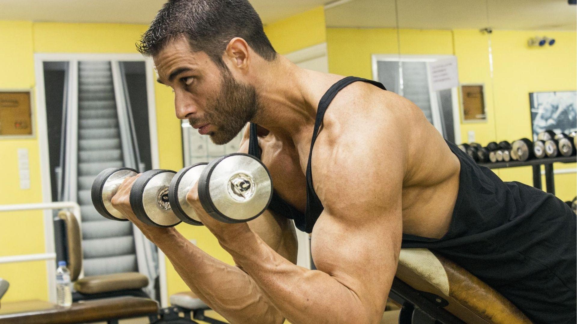 Fit man in tank top performs heavy dumbbell spider curls on incline bench, biceps peak contraction shown.